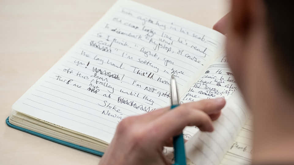 paper 桌布 壁紙 — 一隻手用藍色筆在木桌上的橫線筆記本上寫著亂糟糟的草書的特寫。