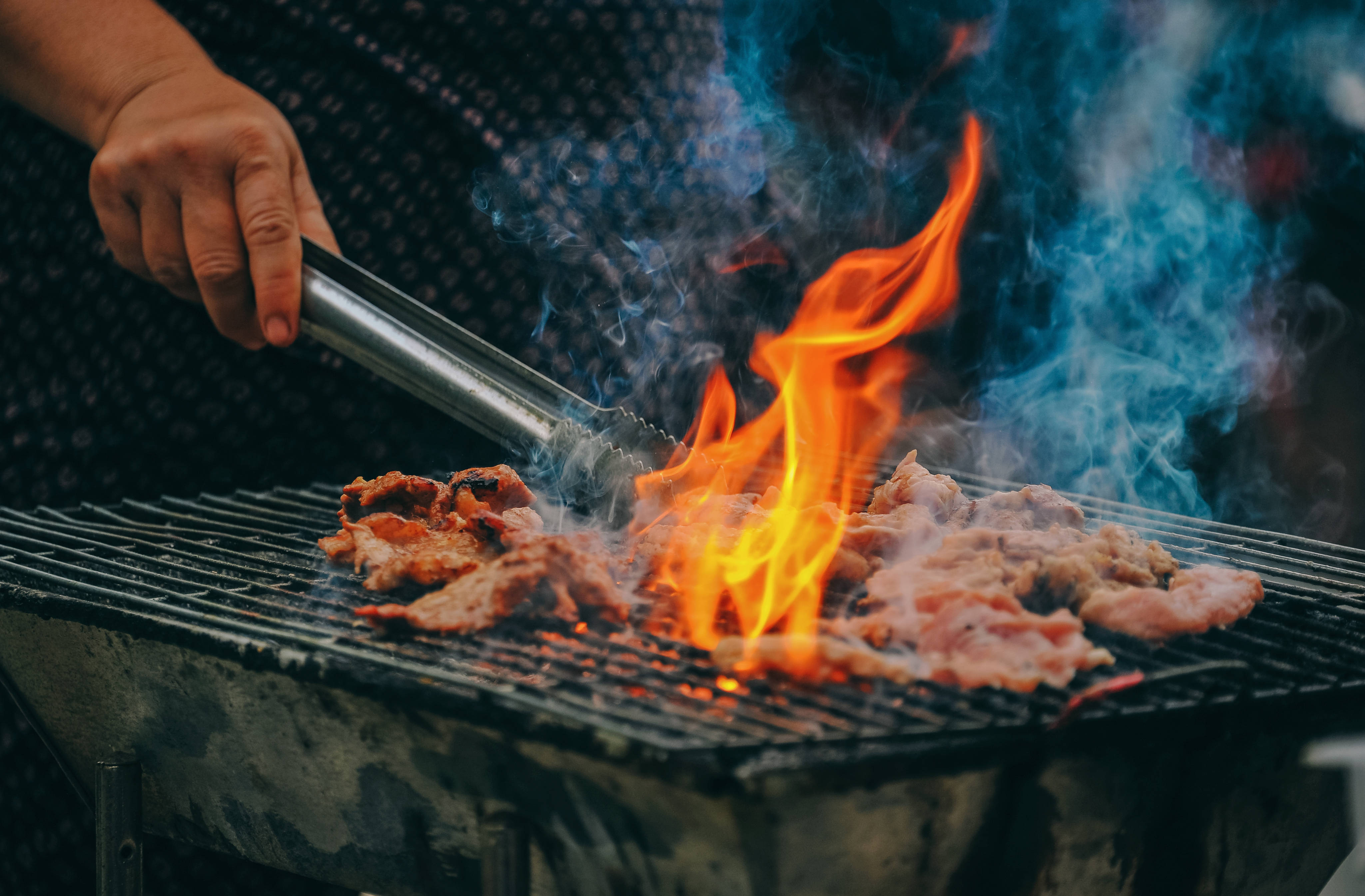 meat 桌布 壁紙 — 一隻手用鉗子將在炭火烤架上滋滋作響的肉翻面，火焰呈鮮豔的橙色，煙霧裊裊升起。