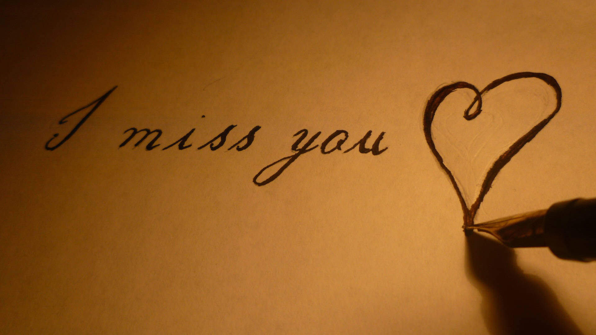 Missing 桌布 壁紙 — 一隻手用鋼筆在心形旁邊的暖色調紙上寫「I miss you」，燈光柔和。