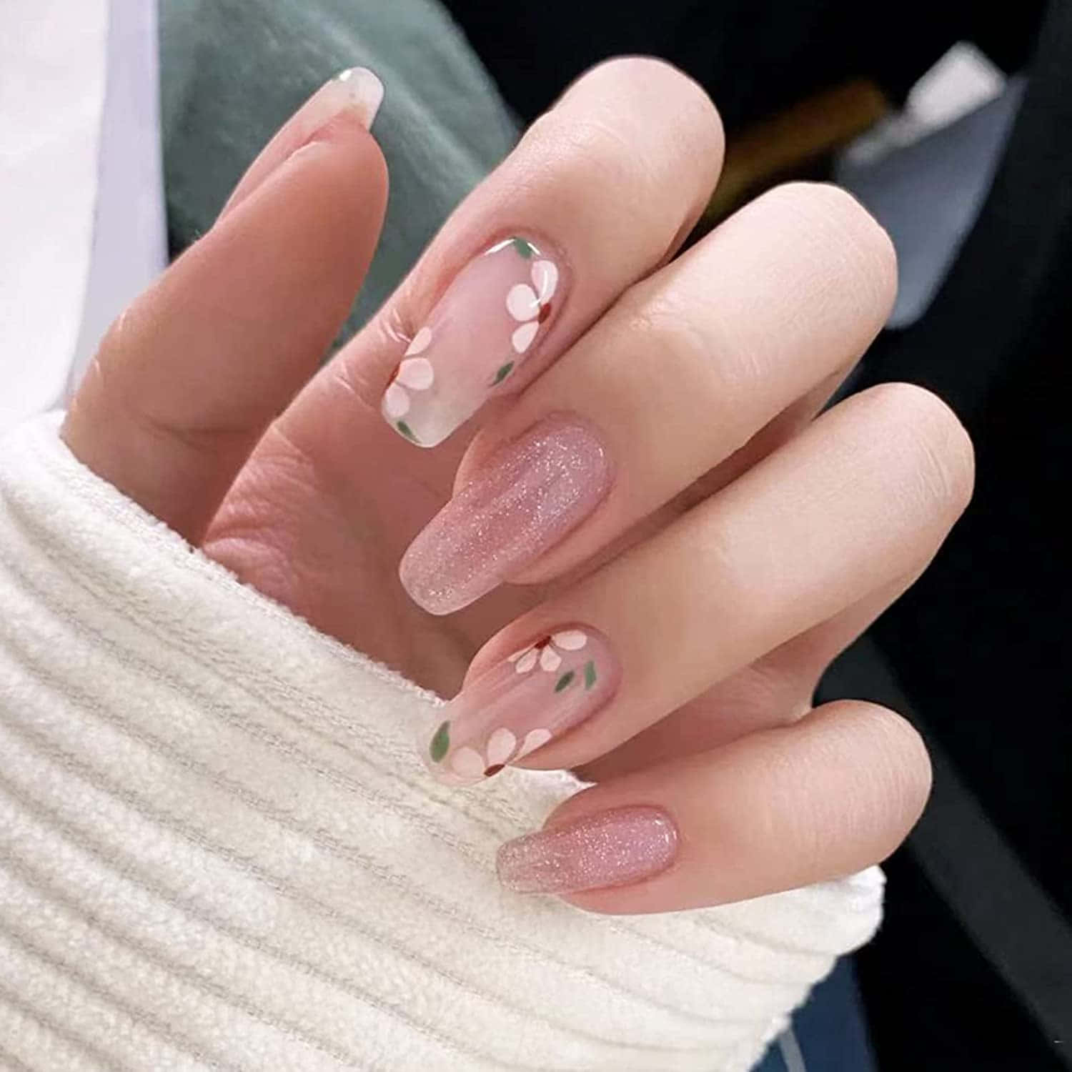 Pink Nail Art 桌布 壁紙 — 一隻手的特寫，帶有精緻的粉紅色閃粉指甲，飾有白色櫻花貼紙，擱在羅紋白色毛衣袖上。