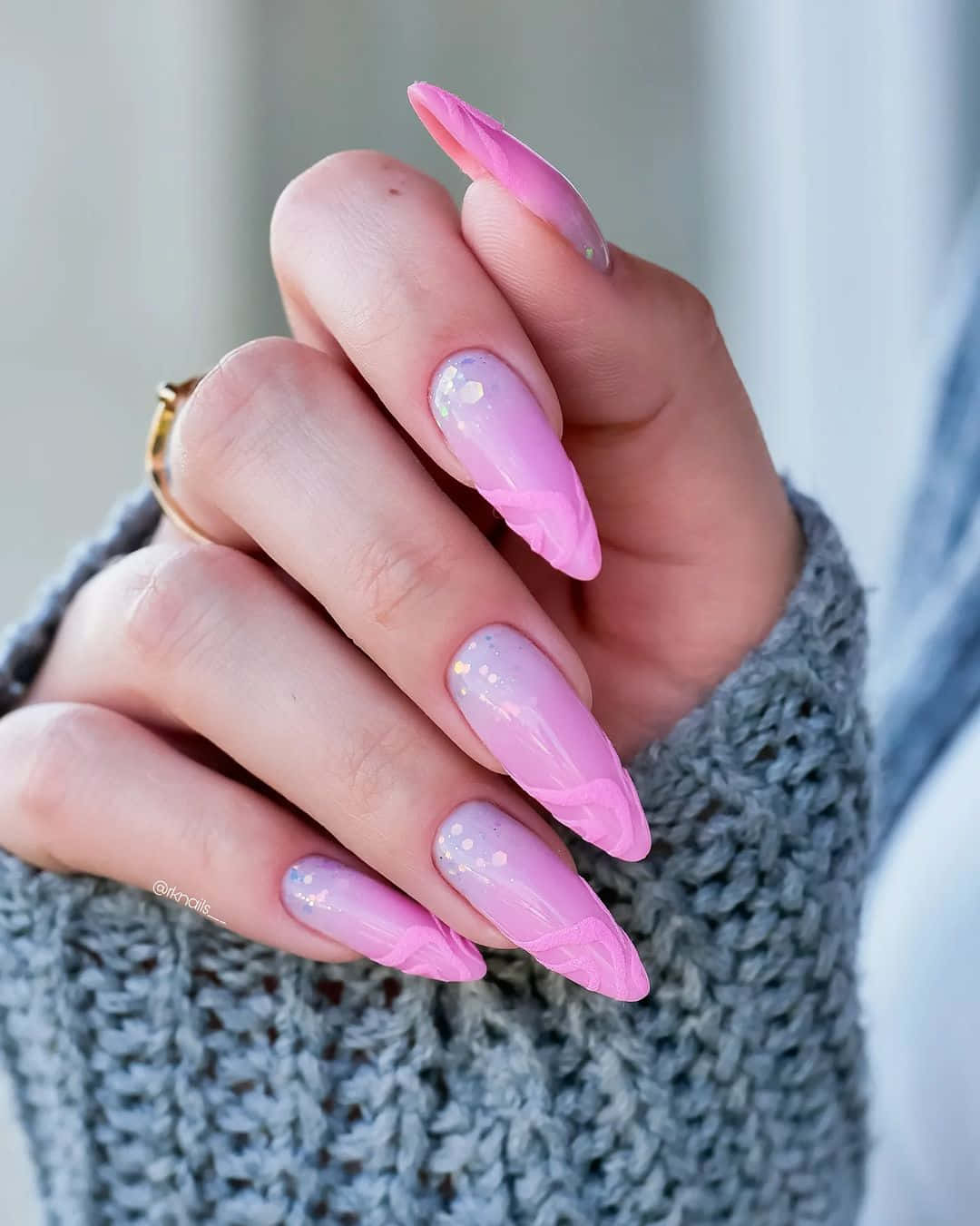Pink Nail Art 桌布 壁紙 — 一隻手的特寫，有長粉紅色亮粉指甲和金戒指，從灰色針織衫袖口露出。