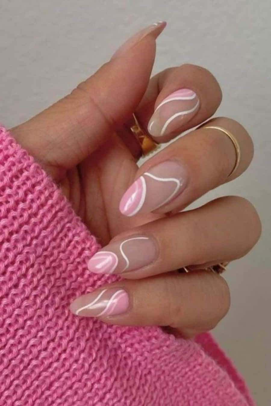 Pink Nail Art 桌布 壁紙 — 一隻手穿著粉紅色針織袖，展示帶有柔和粉紅和白色抽象漩渦及金色戒指的指甲。