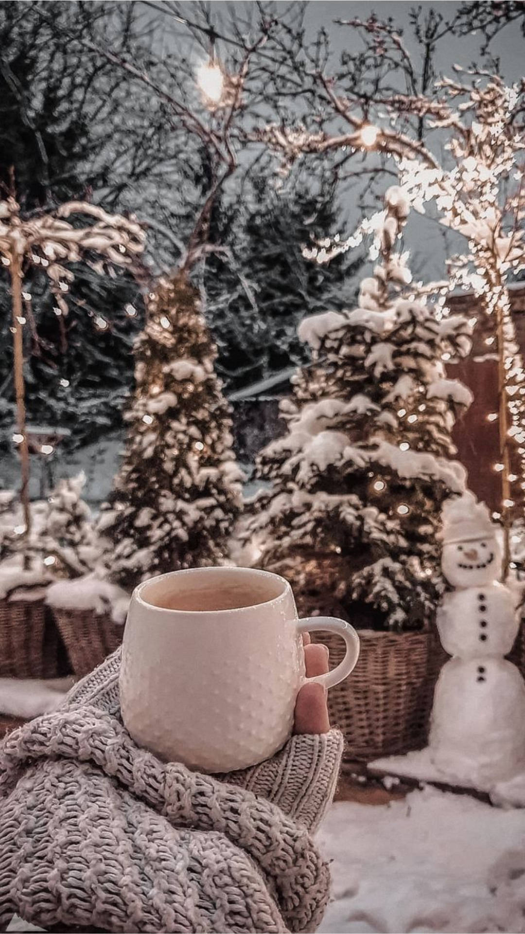 冬天 桌布 壁紙 — 一隻手穿著舒適的針織毛衣，在雪花飄落的庭園背景下，握著一個溫暖的馬克杯，背景有閃爍的燈光、雪人和結霜的樹木。
