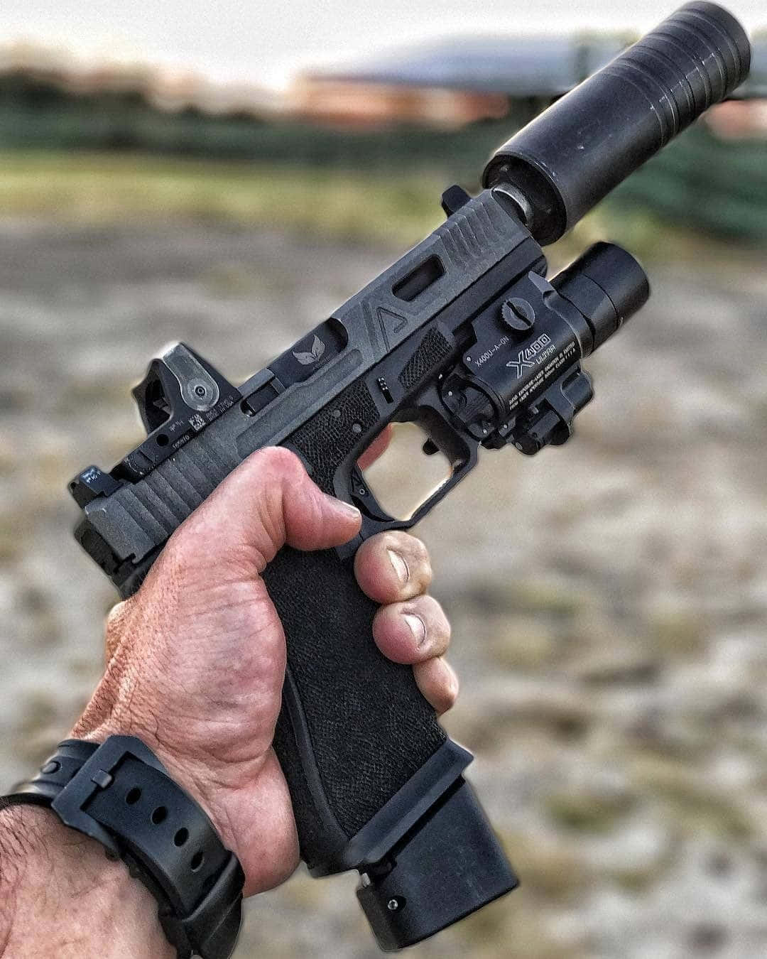 weapon 桌布 壁紙 — 一隻手緊握一支帶有消音器和手電筒的黑色戰術手槍，背景是模糊的戶外環境。