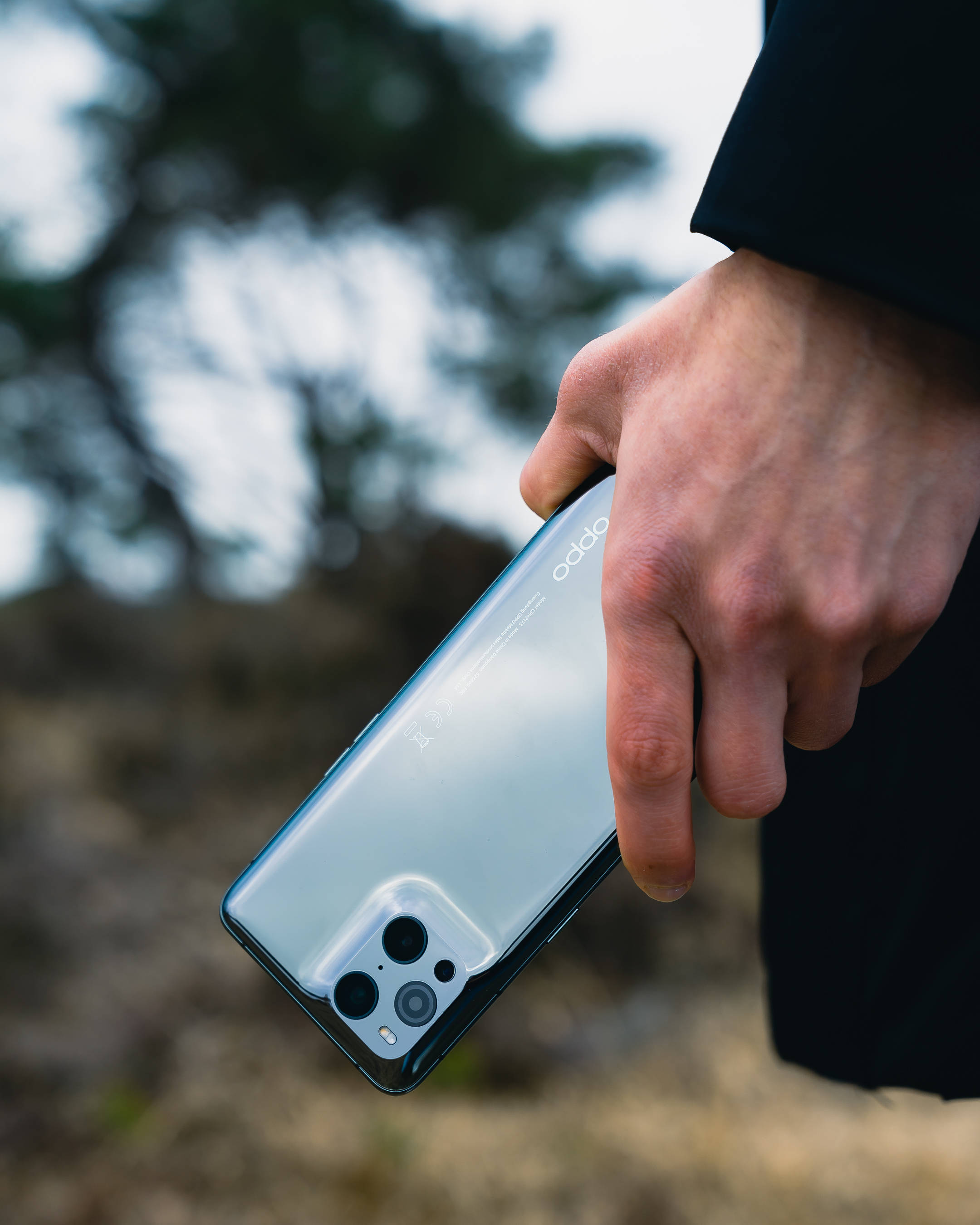 oppo 桌布 壁紙 — 一隻手緊握一款纖薄的銀色 OPPO 智慧型手機，配備四鏡頭設計，背景為模糊的戶外景色。