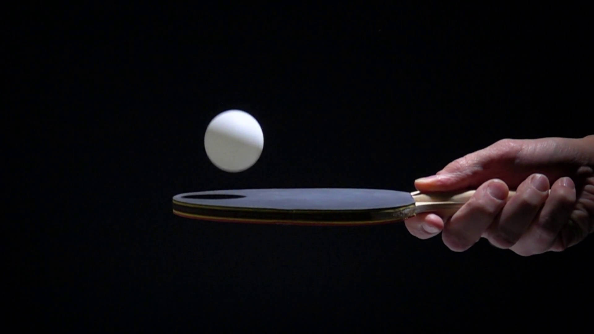 Table Tennis 桌布 壁紙 — 一隻手緊握乒乓球拍，準備擊打懸浮在半空中的白色球，背景為純黑色。