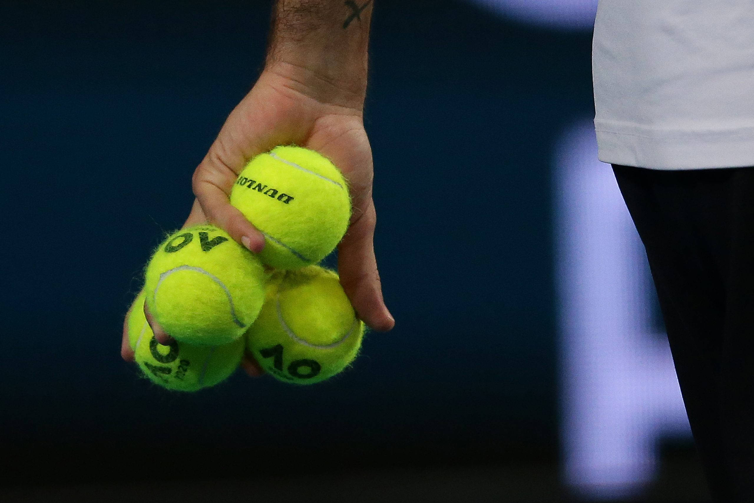 Australian Open 桌布 壁紙 — 一隻手緊握數個鮮黃色網球的特寫，網球上有明顯的品牌標誌，背景為深藍色。