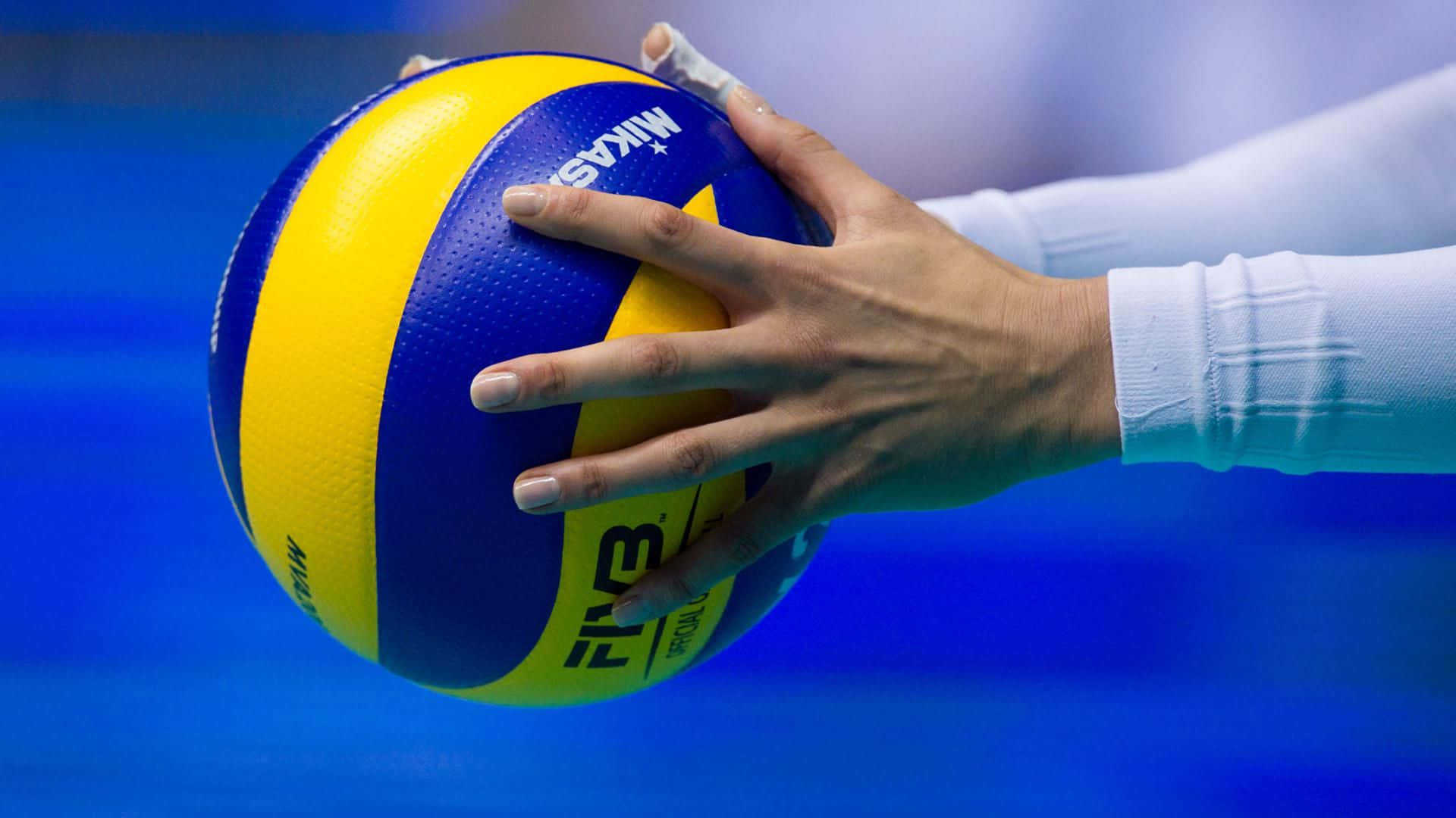 Volleyball 桌布 壁紙 — 一隻手緊握藍黃色 Mikasa 排球的特寫，背景是模糊的藍色球場。