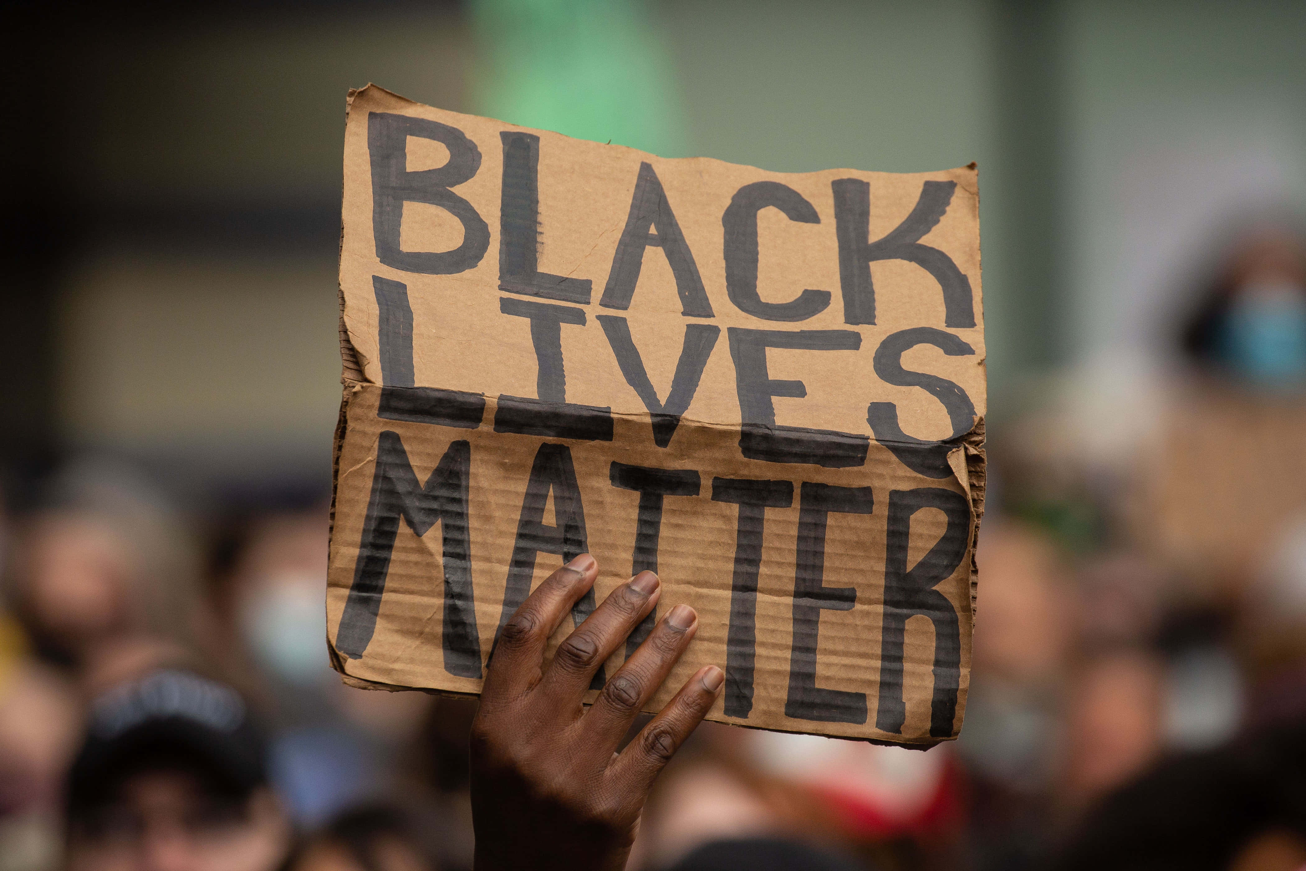 Racism 桌布 壁紙 — 一隻手舉著一個手工製作的紙板標語，上面寫著「BLACK LIVES MATTER」，背景是模糊的示威者。