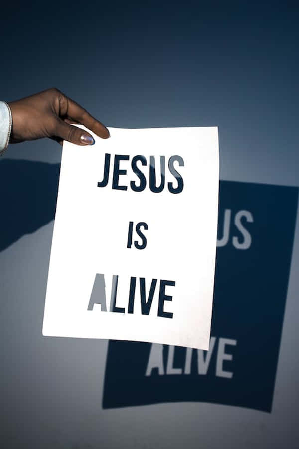 Jesus Name 桌布 壁紙 — 一隻手舉起一個白色標牌，上面有粗體黑色文字寫著「JESUS IS ALIVE」，背景是深藍色漸變。