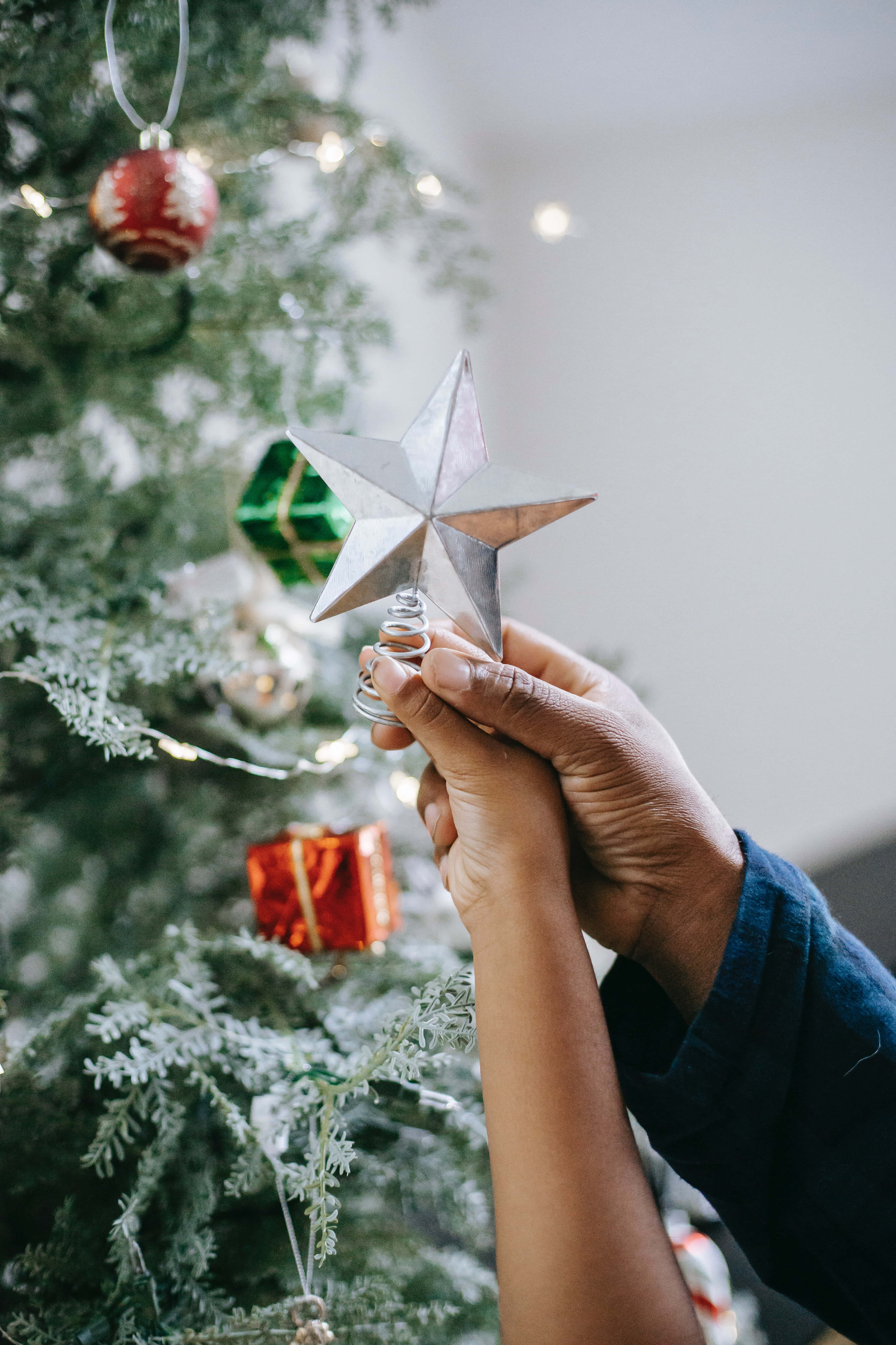 Christmas Star 桌布 壁紙 — 一隻手輕輕地將銀色星星樹頂裝飾放在一棵裝飾著溫暖燈光和彩色飾品的常綠聖誕樹上。