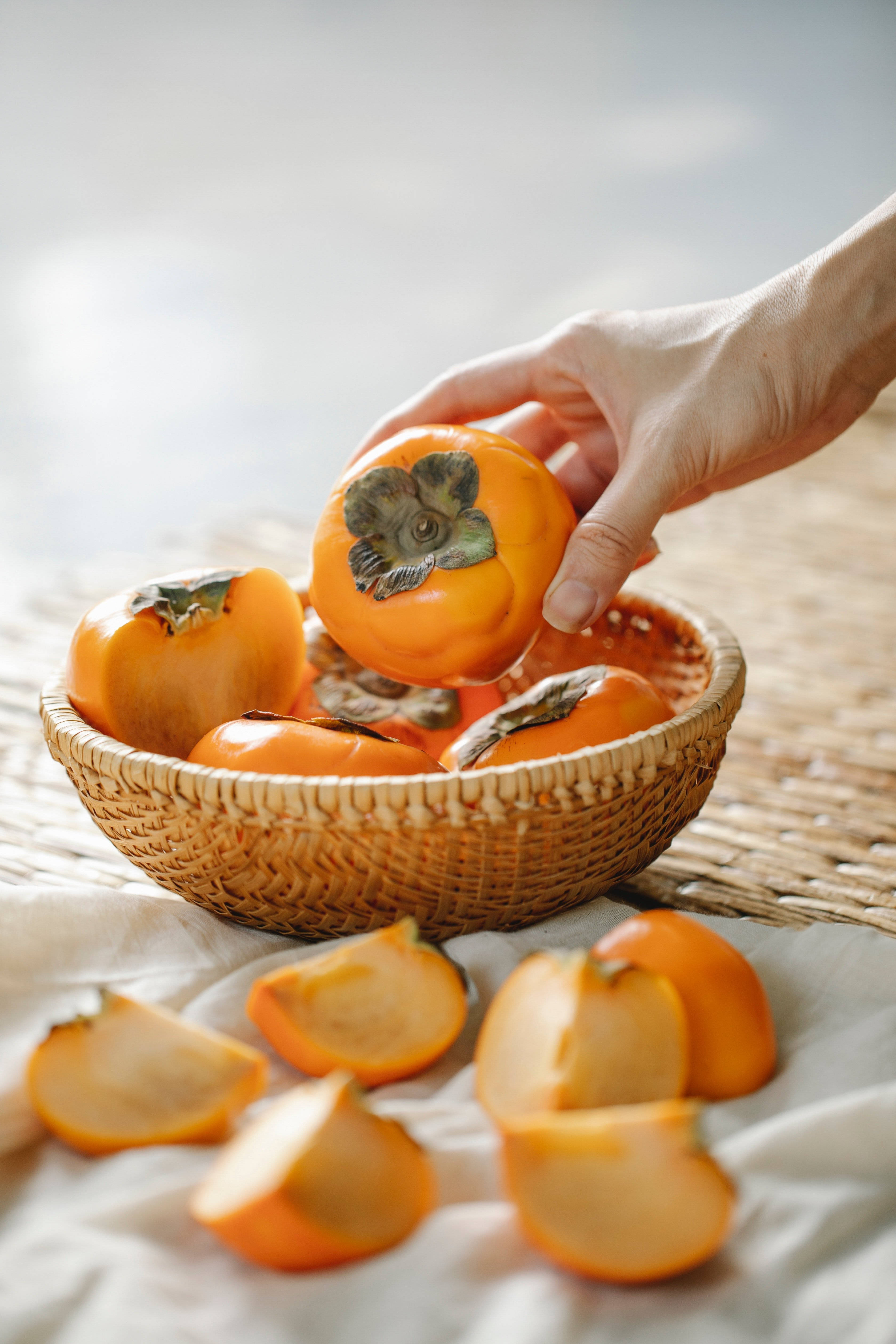 Fruit 桌布 壁紙 — 一隻手輕輕地從一個裝滿鮮豔橙色水果的編織籃子裡拿起一個成熟的柿子，切半的柿子散落在柔軟的白布上。