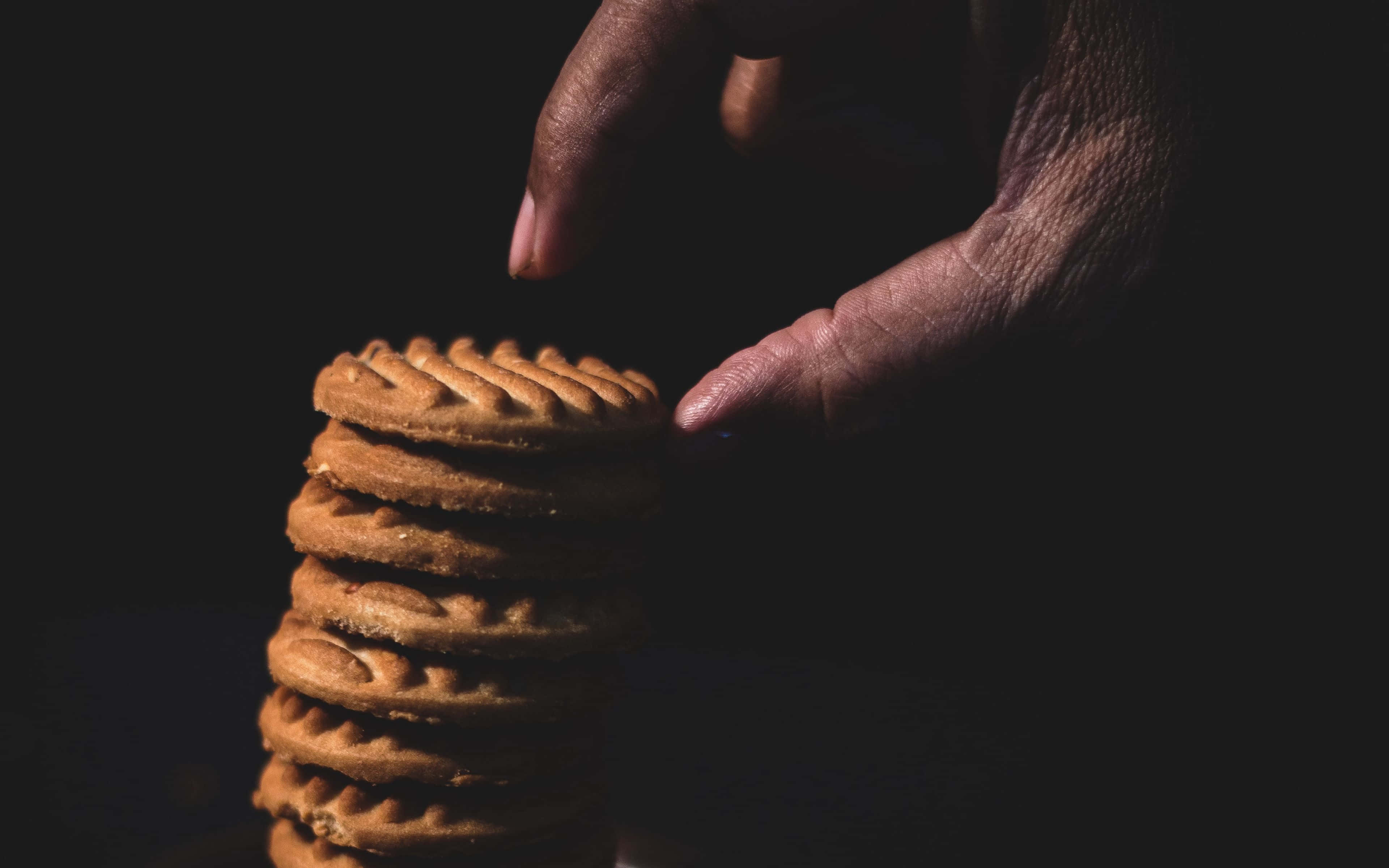 Cookies 桌布 壁紙 — 一隻手輕輕地從深色背景前的高堆中拿起一塊金黃色的鬆餅餅乾。