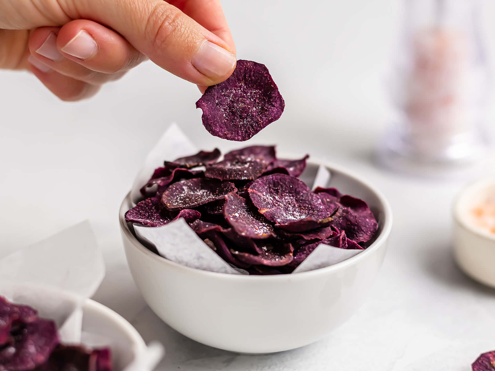Purple Sweet Potato 桌布 壁紙 — 一隻手輕輕地從鋪有烘焙紙的白色碗中拿起一塊閃閃發光的紫色地瓜片。