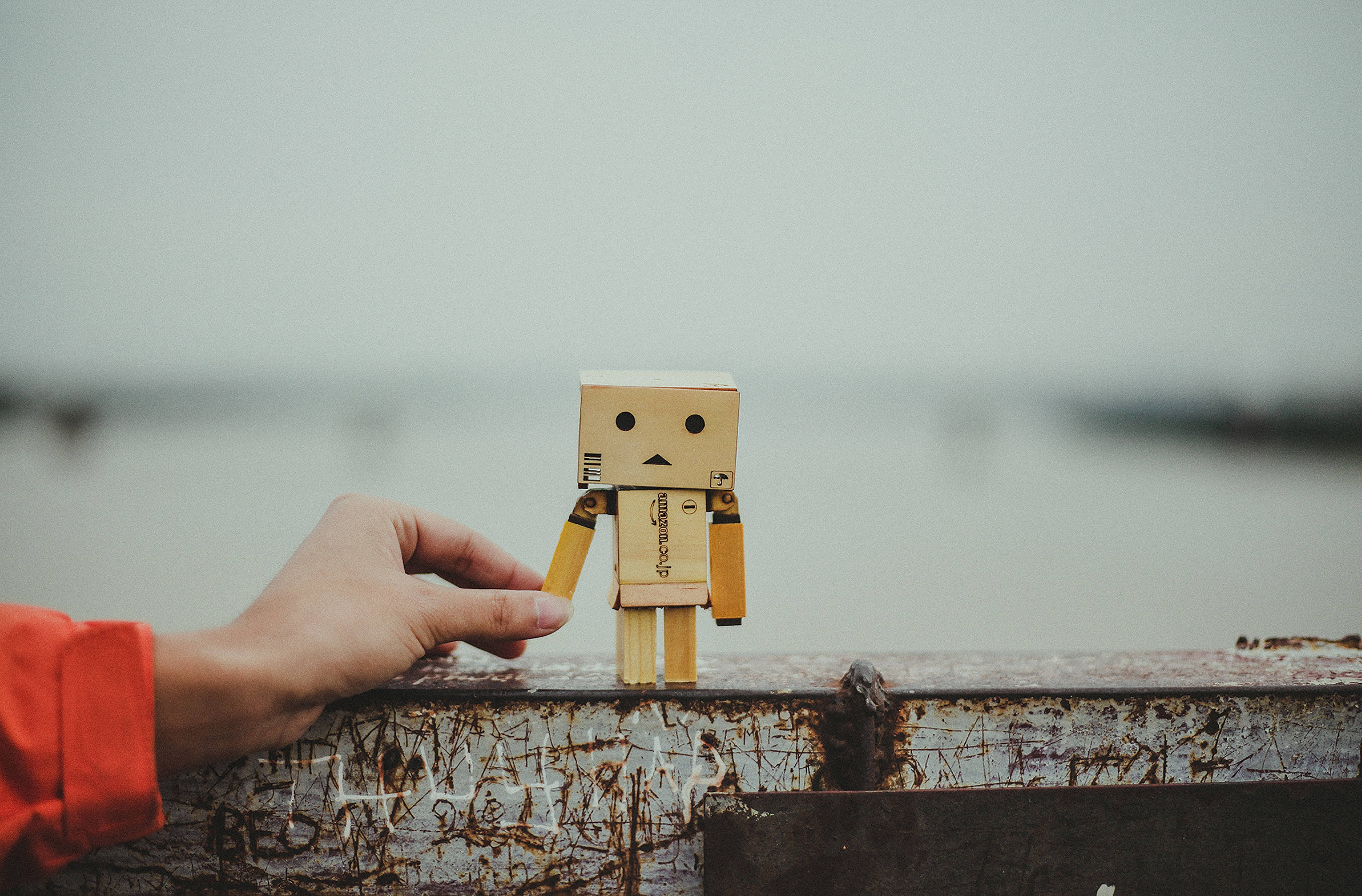Friendship 桌布 壁紙 — 一隻手輕輕地拿著一個小紙板機器人公仔，放在生鏽的金屬欄杆上，俯瞰模糊的水景。