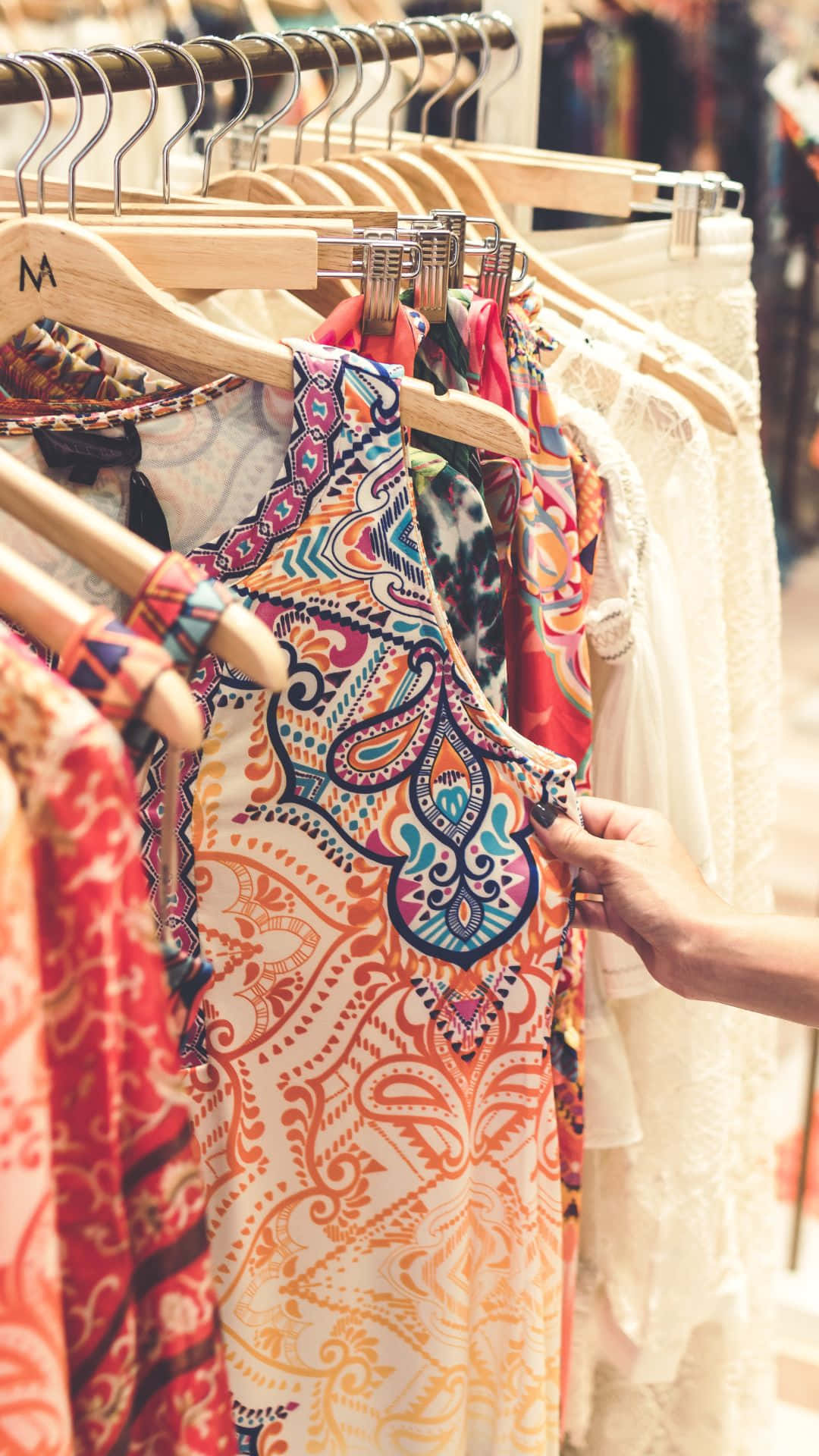 Clothes 桌布 壁紙 — 一隻手輕輕地拿著掛在木衣架上、色彩鮮豔的佩斯利圖案連衣裙，背景是精品店內色彩繽紛的衣架。