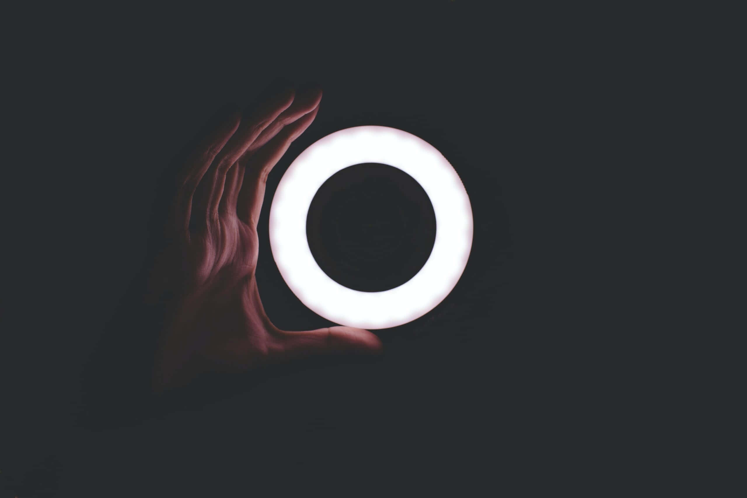 Ring Light 桌布 壁紙 — 一隻手輕輕托著一個發光的白色戒指，背景為深色，投射出柔和陰影，營造出簡約而未來感的氛圍。