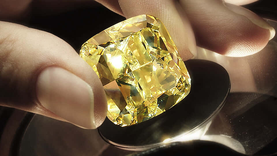 Yellow Diamond 桌布 壁紙 — 一隻手輕輕握住一顆閃耀的黃色枕形切割鑽石，在聚焦光線下於深色反光表面上發光。