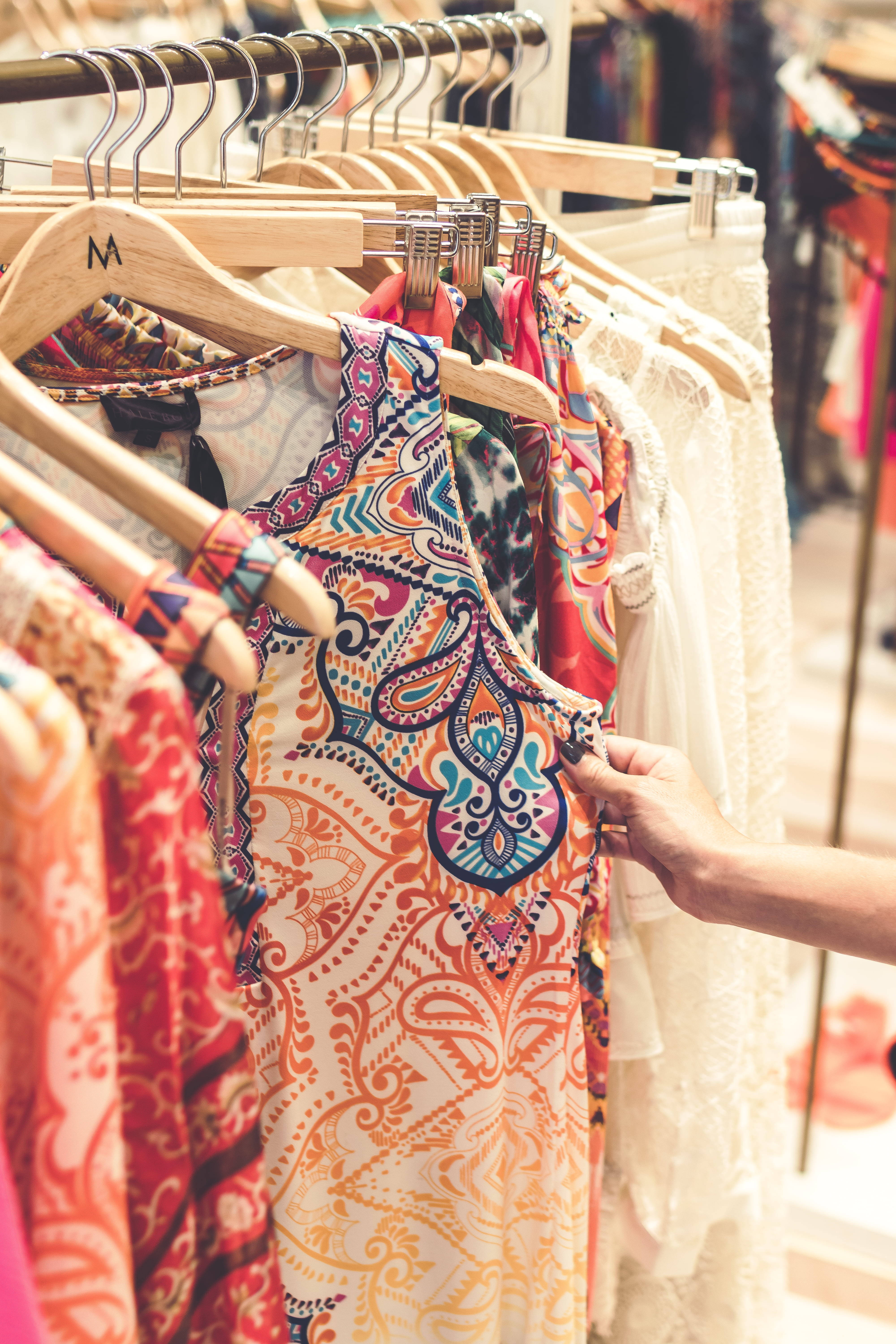 Clothes 桌布 壁紙 — 一隻手輕輕觸摸掛在木衣架上、充滿活力的佩斯利花紋上衣，周圍是精品店裡色彩繽紛的衣物。