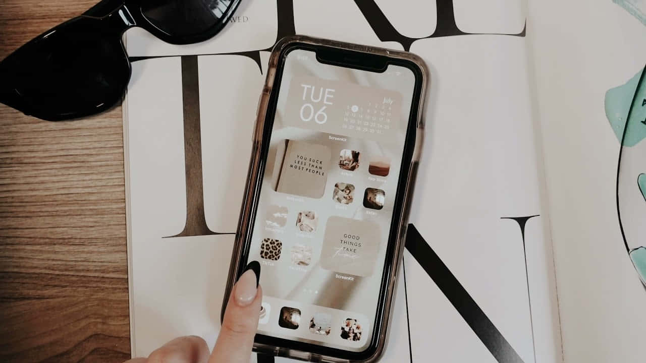 iPhone 美學 桌布 壁紙 — 一隻手輕點一部智慧型手機，螢幕上顯示一個簡約風格的桌布，包含日曆和應用程式圖示，手機放在木桌上的一本雜誌上。