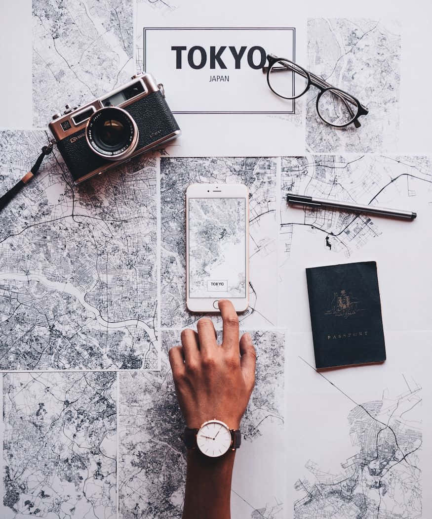 Passport 桌布 壁紙 — 一隻手輕點顯示東京地圖的手機，周圍是旅行裝備，背景為城市地圖。