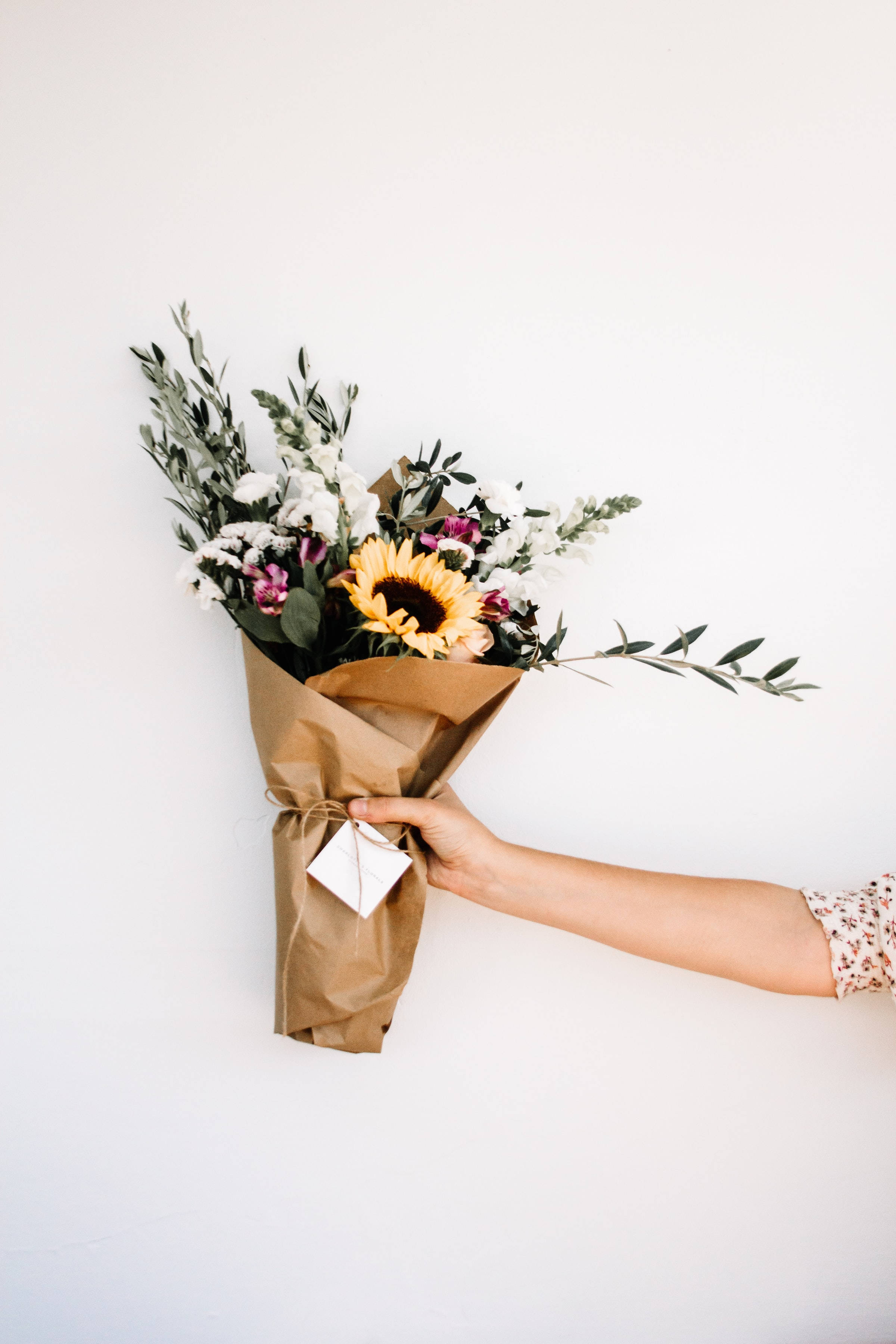 Bouquet 桌布 壁紙 — 一隻手遞出一個質樸的棕色紙花束，包含一朵鮮豔的向日葵、白色花朵和綠葉，背景是純白牆壁。