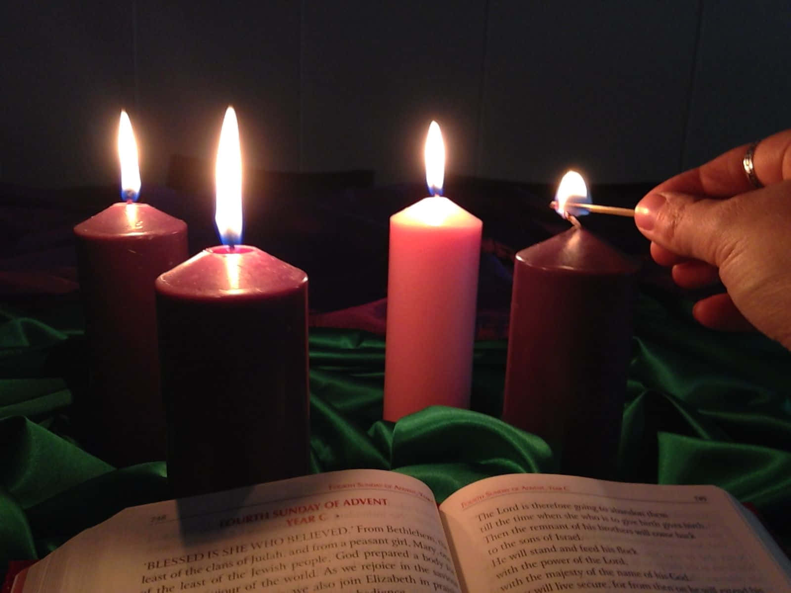 Advent 桌布 壁紙 — 一隻手點燃一支紫色降臨期蠟燭，旁邊有三支其他蠟燭放在綠色布上，下方有一本打開的聖經。
