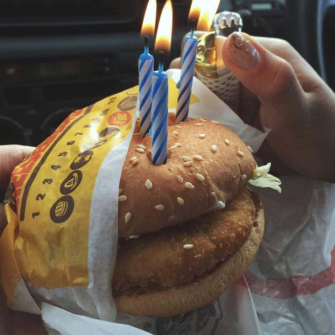 fast food 桌布 壁紙 — 一隻手點燃插在芝麻包漢堡上的藍色條紋生日蠟燭，漢堡放在有品牌標誌的包裝紙中。