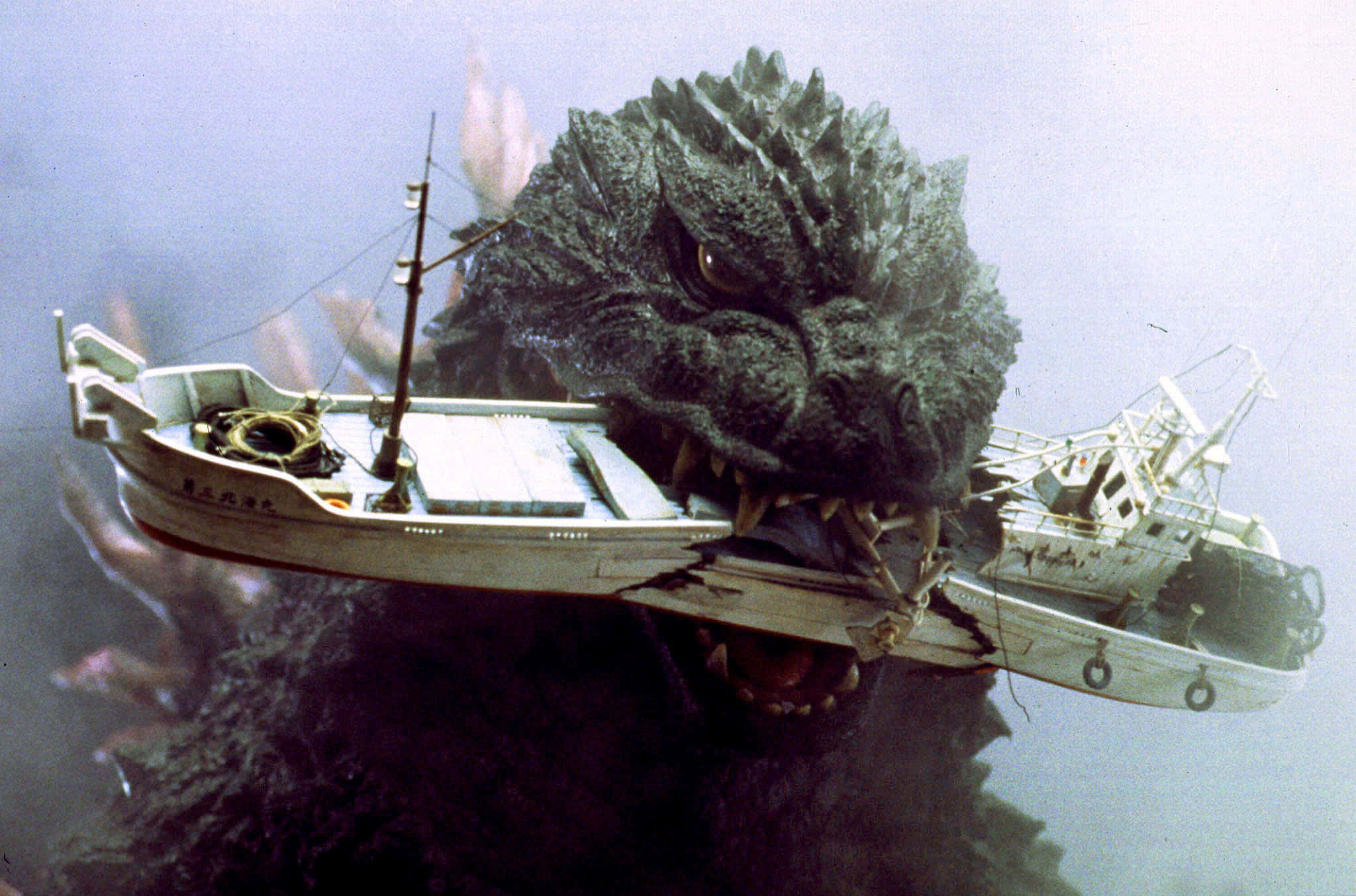 Godzilla 2000 桌布 壁紙 — 一隻擁有尖刺灰色皮膚的怪獸哥吉拉在渾濁的海水中咬住一艘小漁船。