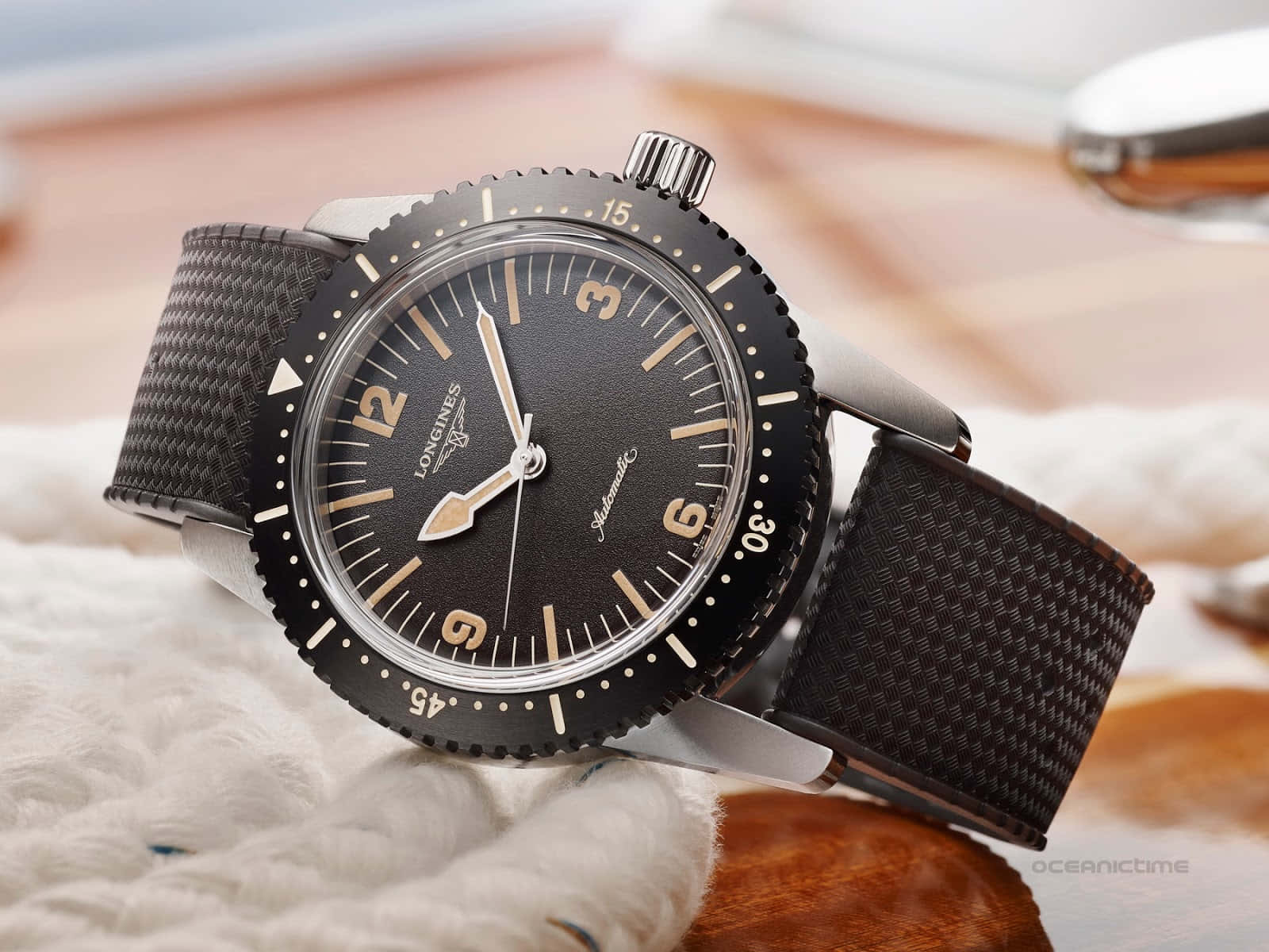 圖片 桌布 壁紙 — 一隻時尚的黑色 Longines 手錶，配有紋理錶帶，放在柔軟的白色布料上，背景是溫暖的木質。