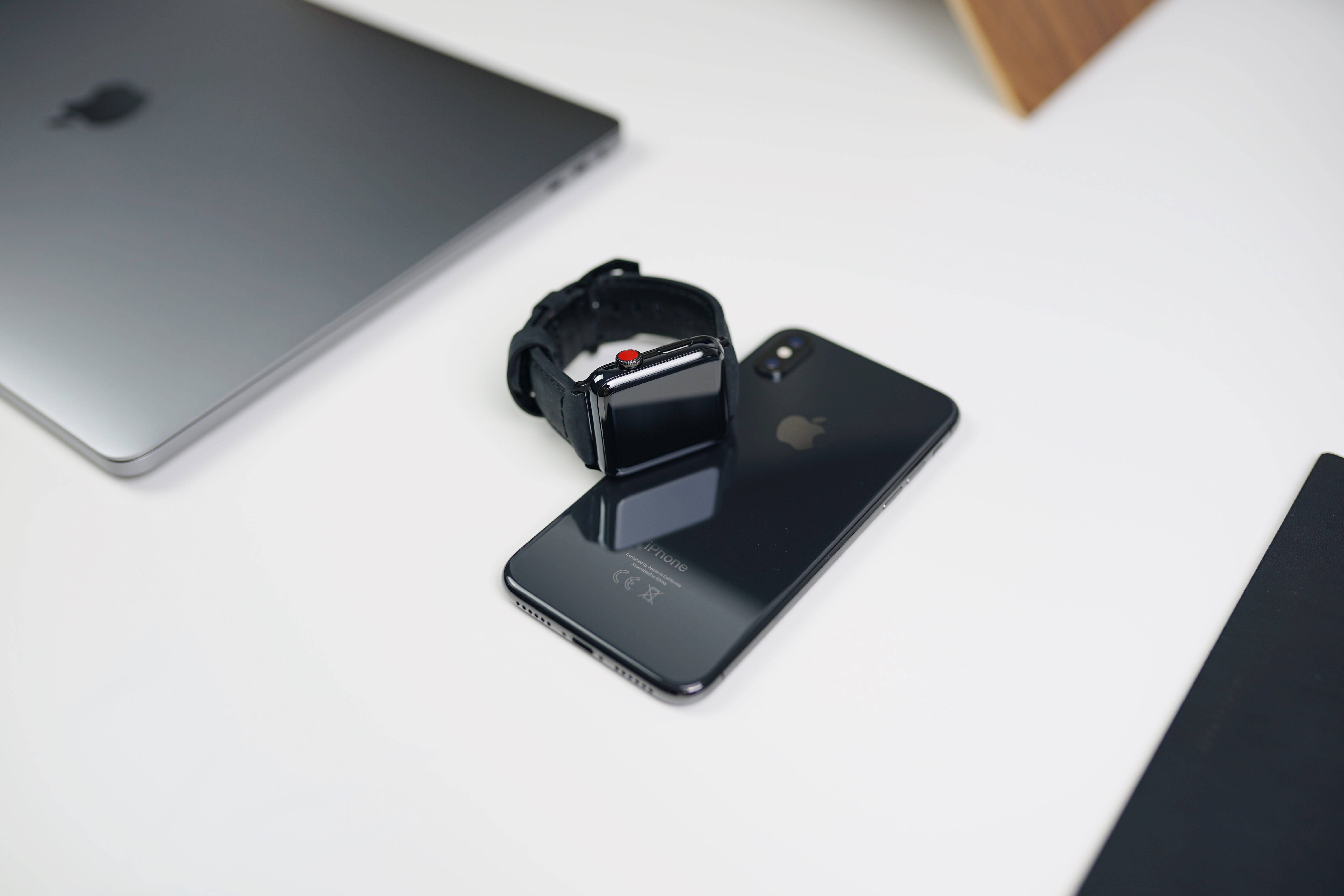 Apple Watch 動態 桌布 壁紙 — 一隻時尚的黑色Apple Watch放在一部相配的iPhone上，置於簡約白色桌子上，旁邊有一部銀色筆記型電腦。