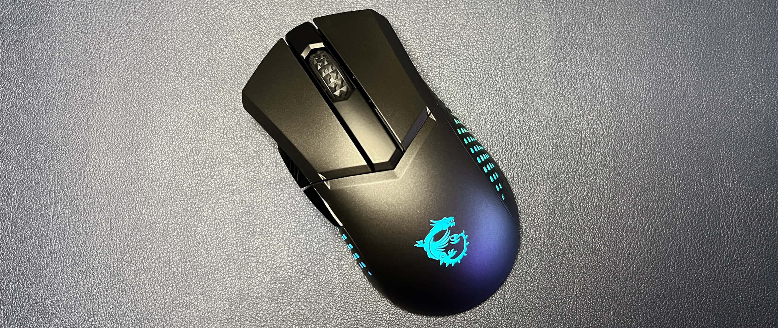 msi 2560x1080 桌布 壁紙 — 一隻時尚的黑色電競滑鼠帶有發光的藍色龍標誌，放置在紋理灰色表面上。