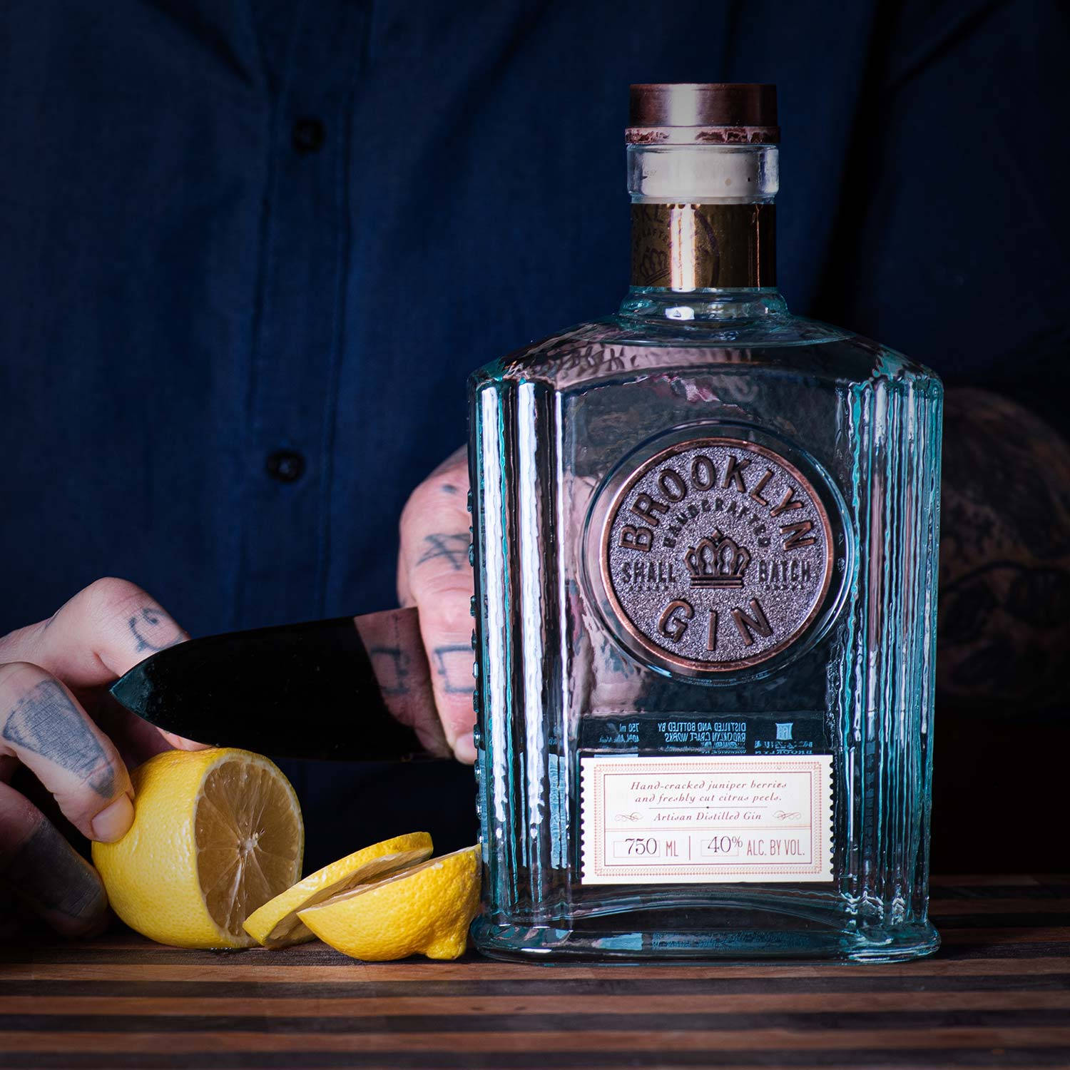 Drink 桌布 壁紙 — 一隻有刺青的手在木桌上，於一瓶透明的 Brooklyn Gin 旁邊切檸檬。