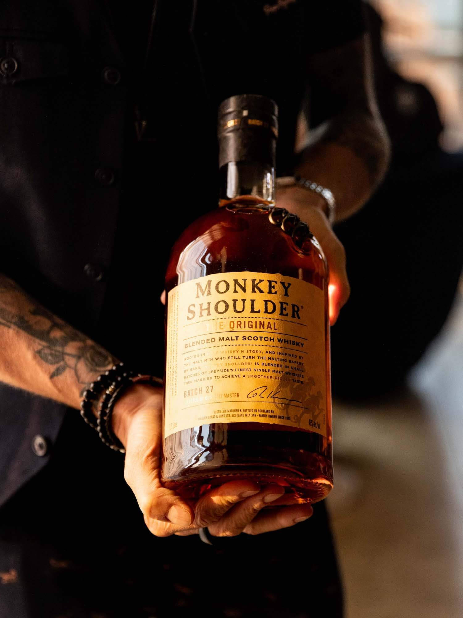 alcohol 桌布 壁紙 — 一隻有刺青的手拿著一瓶Monkey Shoulder威士忌，酒液呈溫暖琥珀色，金色標籤，背景昏暗模糊。