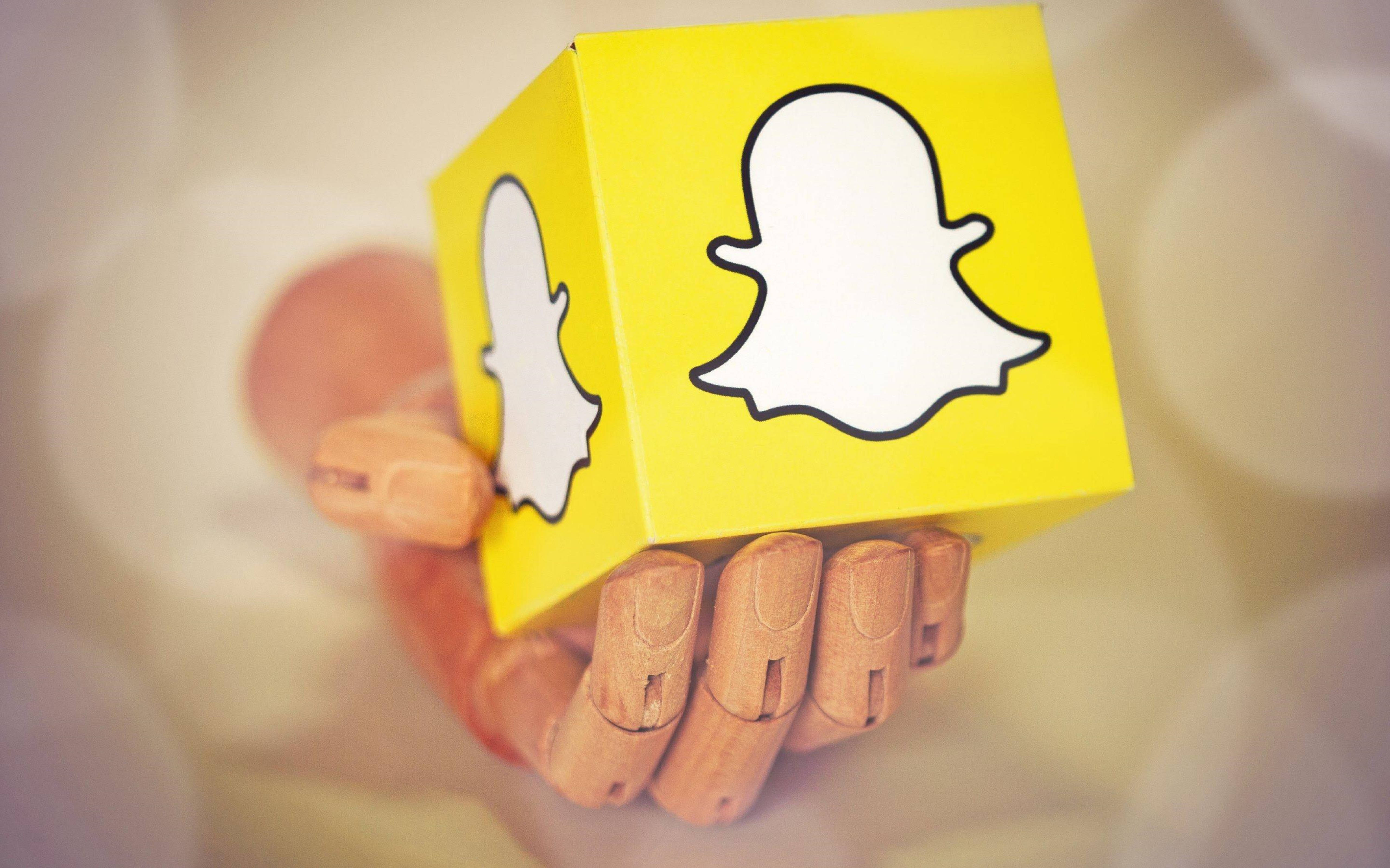 Snapchat 桌布 壁紙 — 一隻木手拿著一個亮黃色立方體，上面有Snapchat鬼魂標誌，背景柔和模糊。
