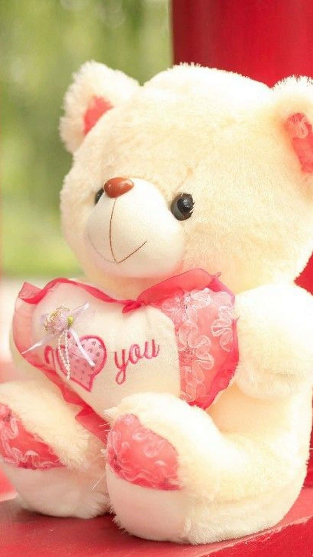 bear love 桌布 壁紙 — 一隻柔軟的奶油色泰迪熊帶有紅色點綴，抱著一個心形枕頭，上面用粉紅色繡著「I love you」，背景是模糊的綠色和紅色。