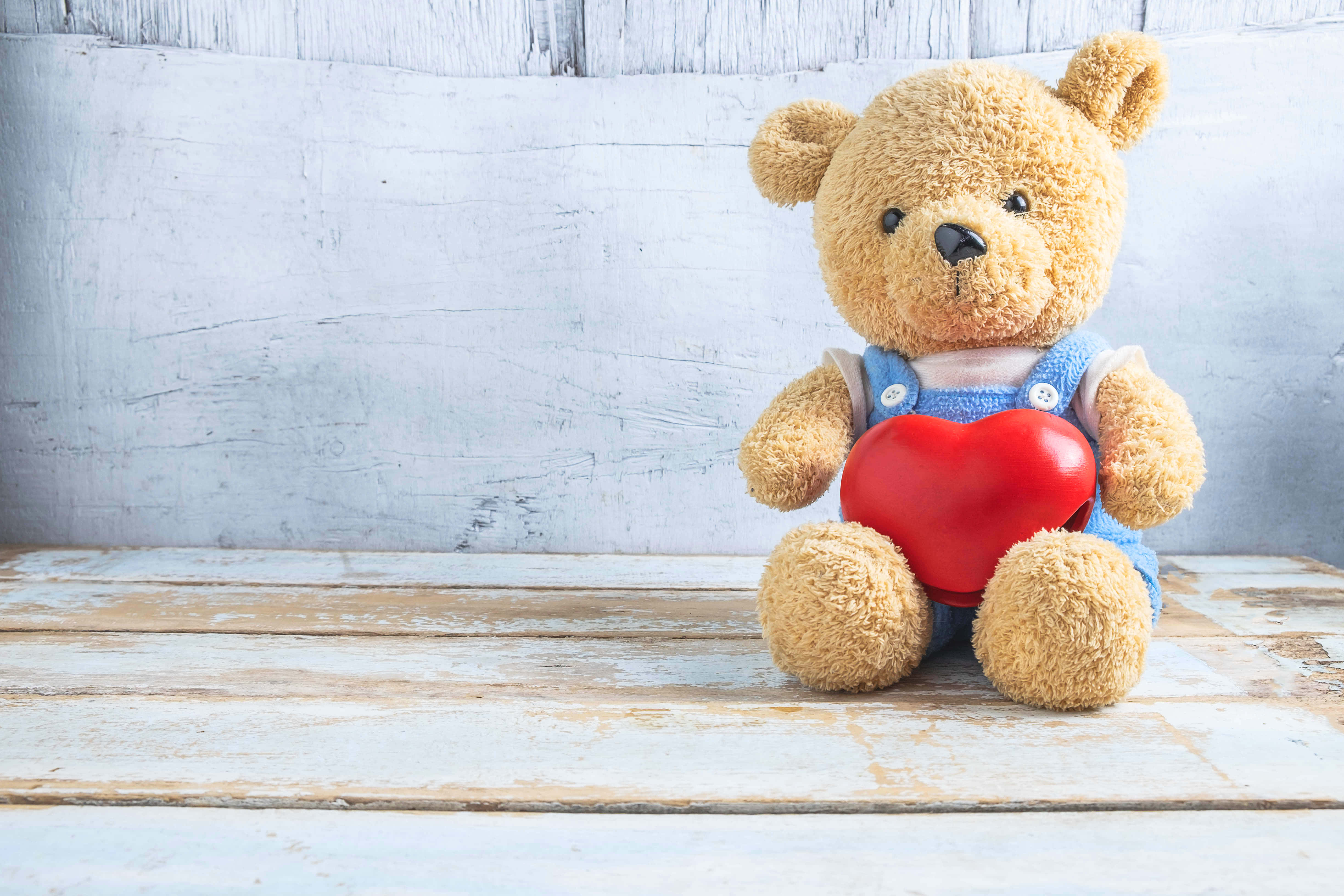 Teddy Bear 桌布 壁紙 — 一隻柔軟的棕色泰迪熊穿著藍色工裝褲，在風化的白色木質背景前拿著一個鮮紅色的心形。