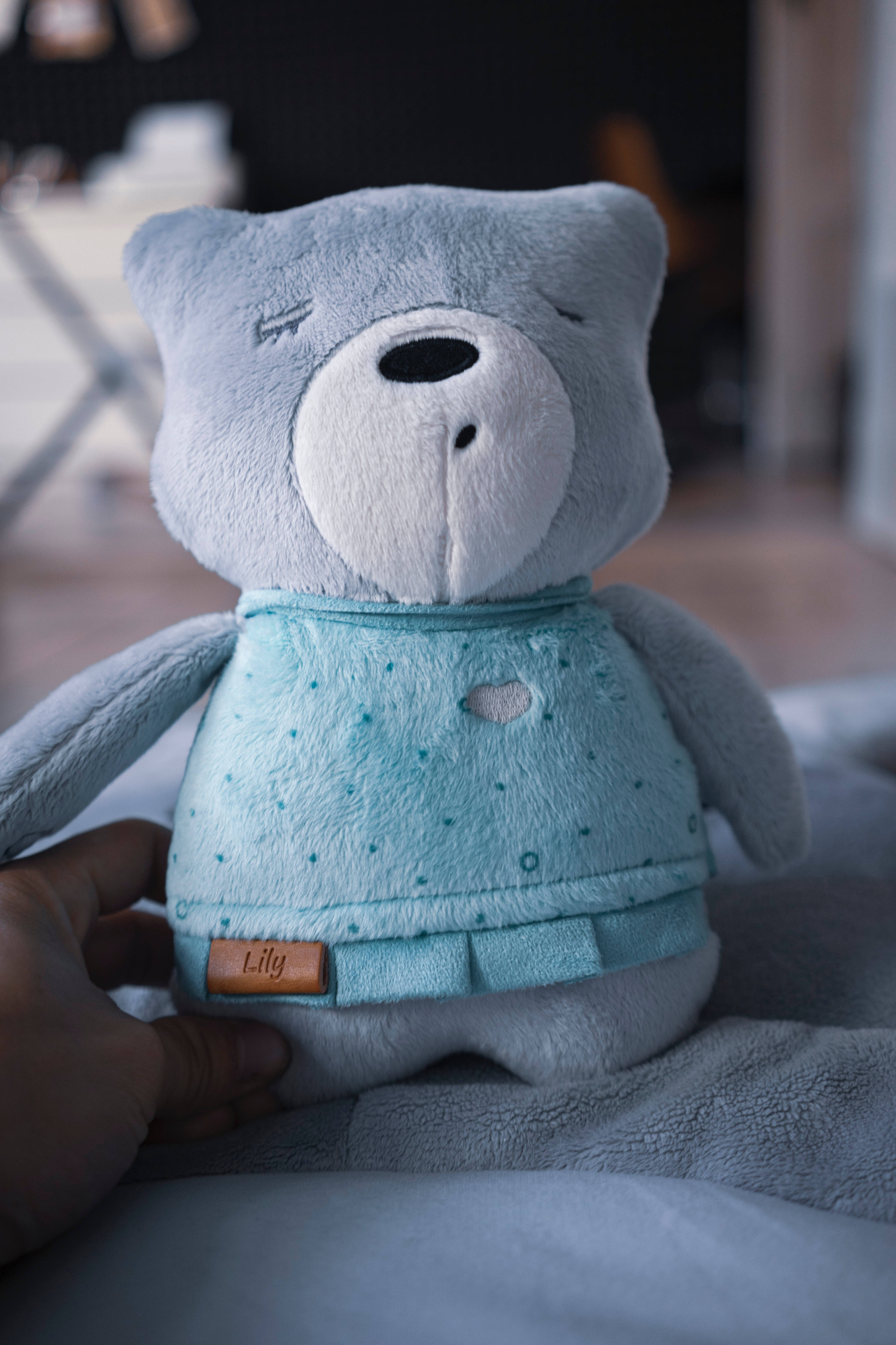 Cute Blue 桌布 壁紙 — 一隻柔軟的淺藍色泰迪熊穿著配套的圓點睡衣套裝，帶有心形補丁和名牌，在舒適的室內環境中被輕輕抱著。