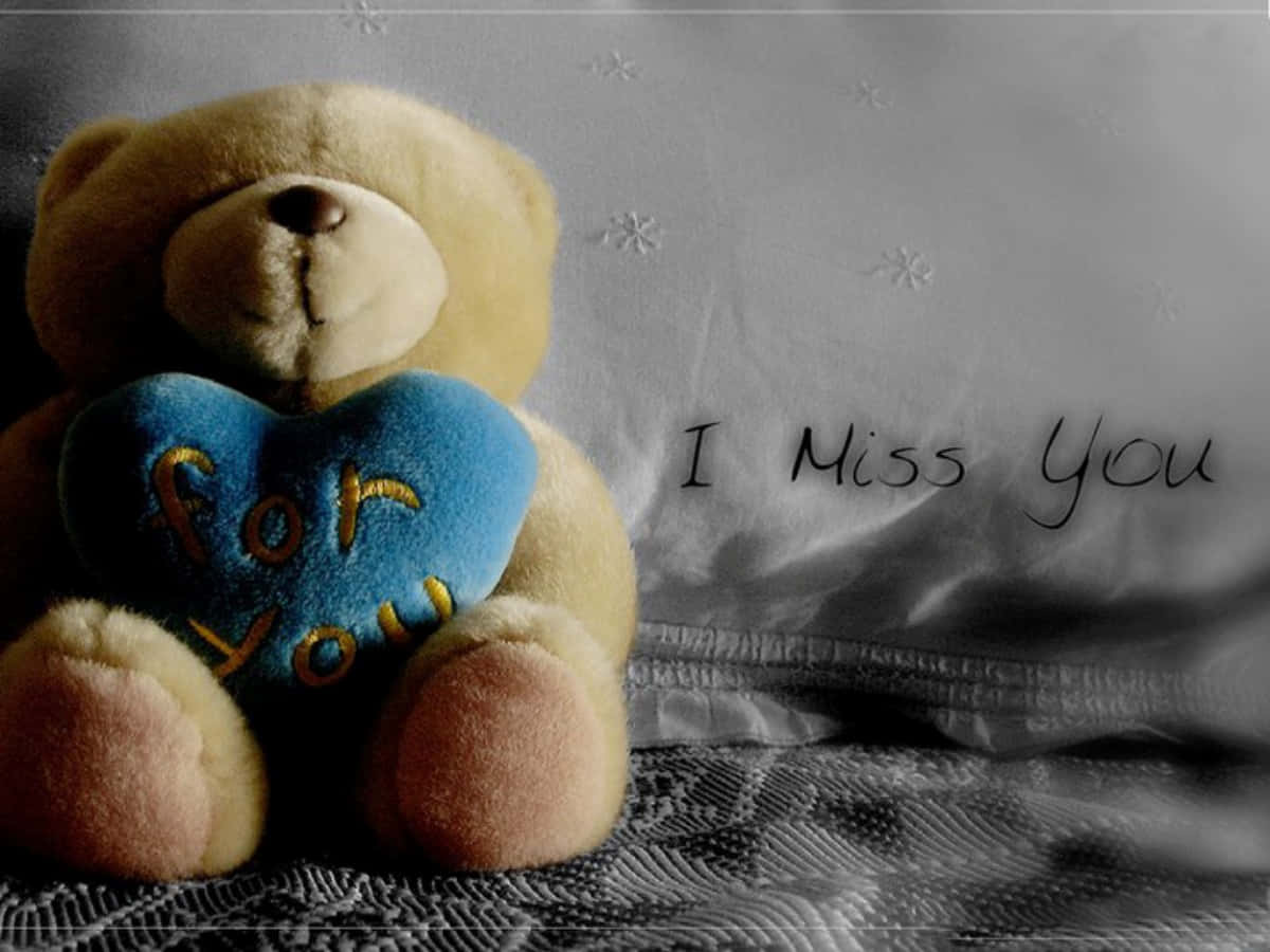 Cute Teddy 桌布 壁紙 — 一隻柔軟的米色泰迪熊抱著一個帶有金色字樣的藍色心形枕頭，坐在有紋理的床罩上，旁邊有手寫文字「我想你」。
