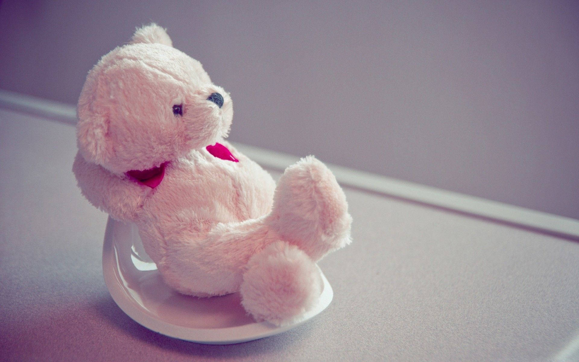 Teddy Bear 桌布 壁紙 — 一隻柔軟的粉紅色泰迪熊繫著紅色蝴蝶結，獨自坐在白色碟子上，背景是柔和的紫色牆壁。
