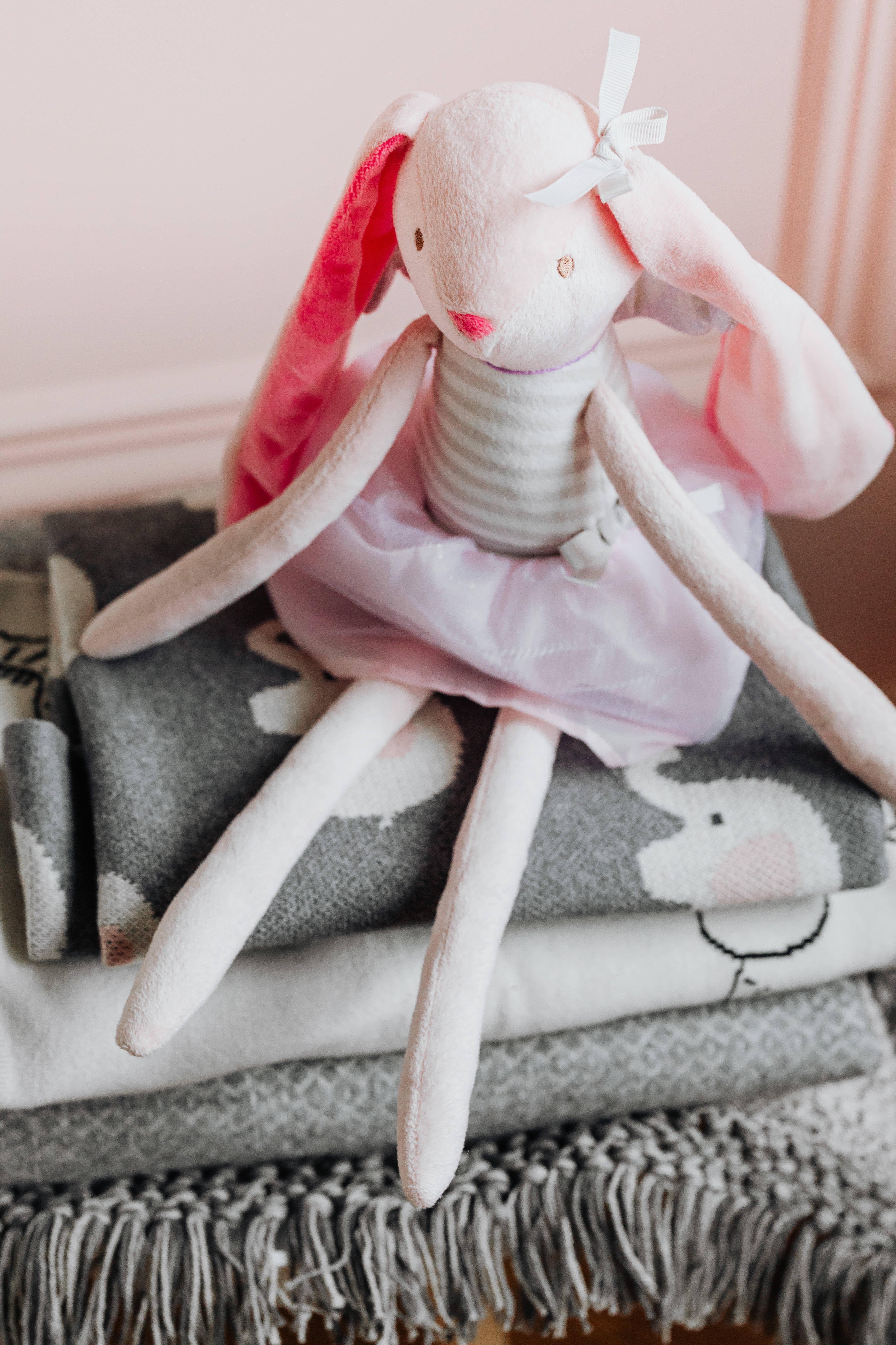 Newborn 桌布 壁紙 — 一隻柔軟的粉紅色長耳絨毛兔，繫著白色蝴蝶結，坐在一堆帶有兔子圖案的舒適灰色和白色毯子上。