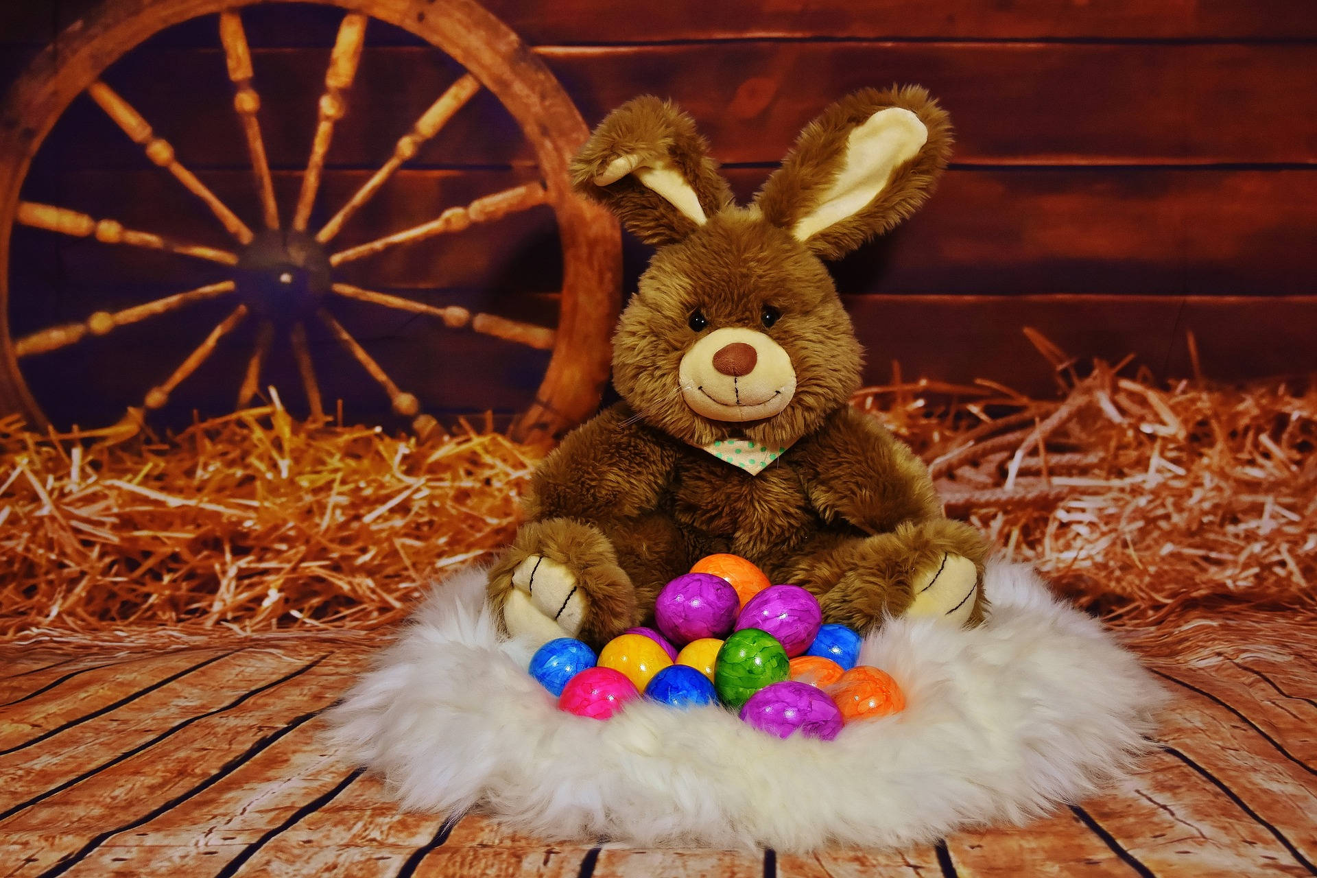 Cute Easter 桌布 壁紙 — 一隻棕色毛絨兔子坐在白色毛茸茸的巢上，拿著色彩繽紛的塑膠復活蛋，背景有木製馬車輪和乾草。