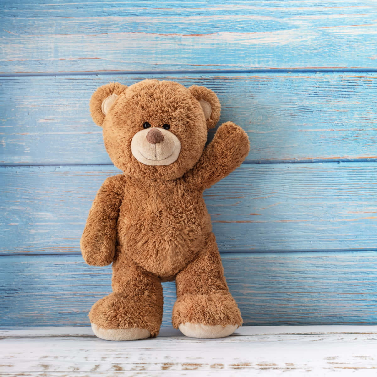 Teddy Bear 桌布 壁紙 — 一隻棕色毛絨泰迪熊在白色木地板上，對著一面風化的藍色木牆愉快地揮手。