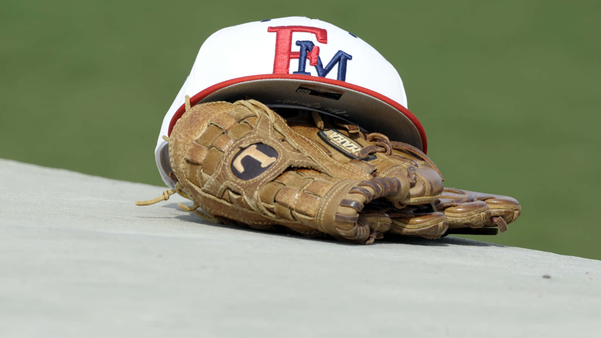 Baseball Gloves 桌布 壁紙 — 一隻棕色皮革棒球手套放在一個陽光明媚的球場上，旁邊是一頂帶有紅藍色「FM」標誌的白色帽子。