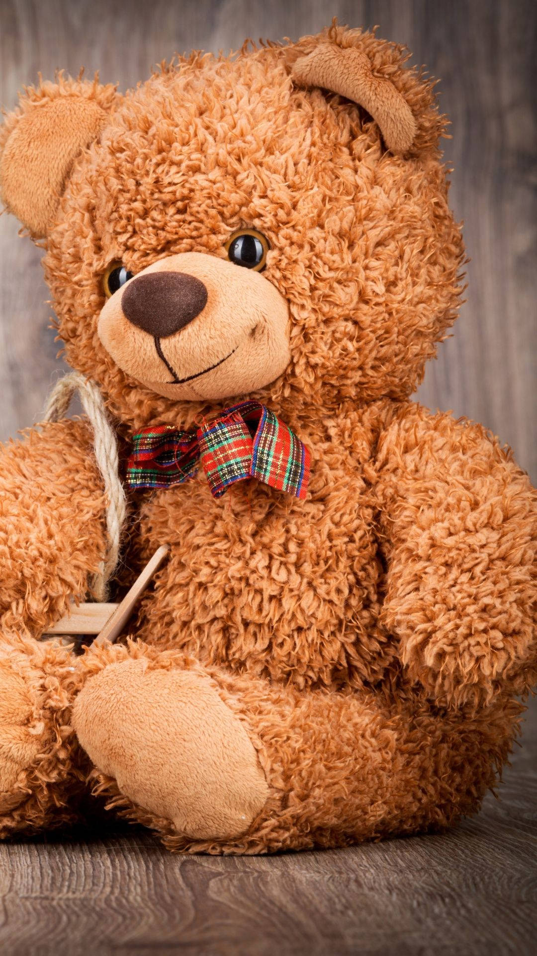 Teddy Bear 桌布 壁紙 — 一隻毛茸茸的棕色泰迪熊，繫著格子蝴蝶結，坐在木質表面上，背景是質樸的木頭。