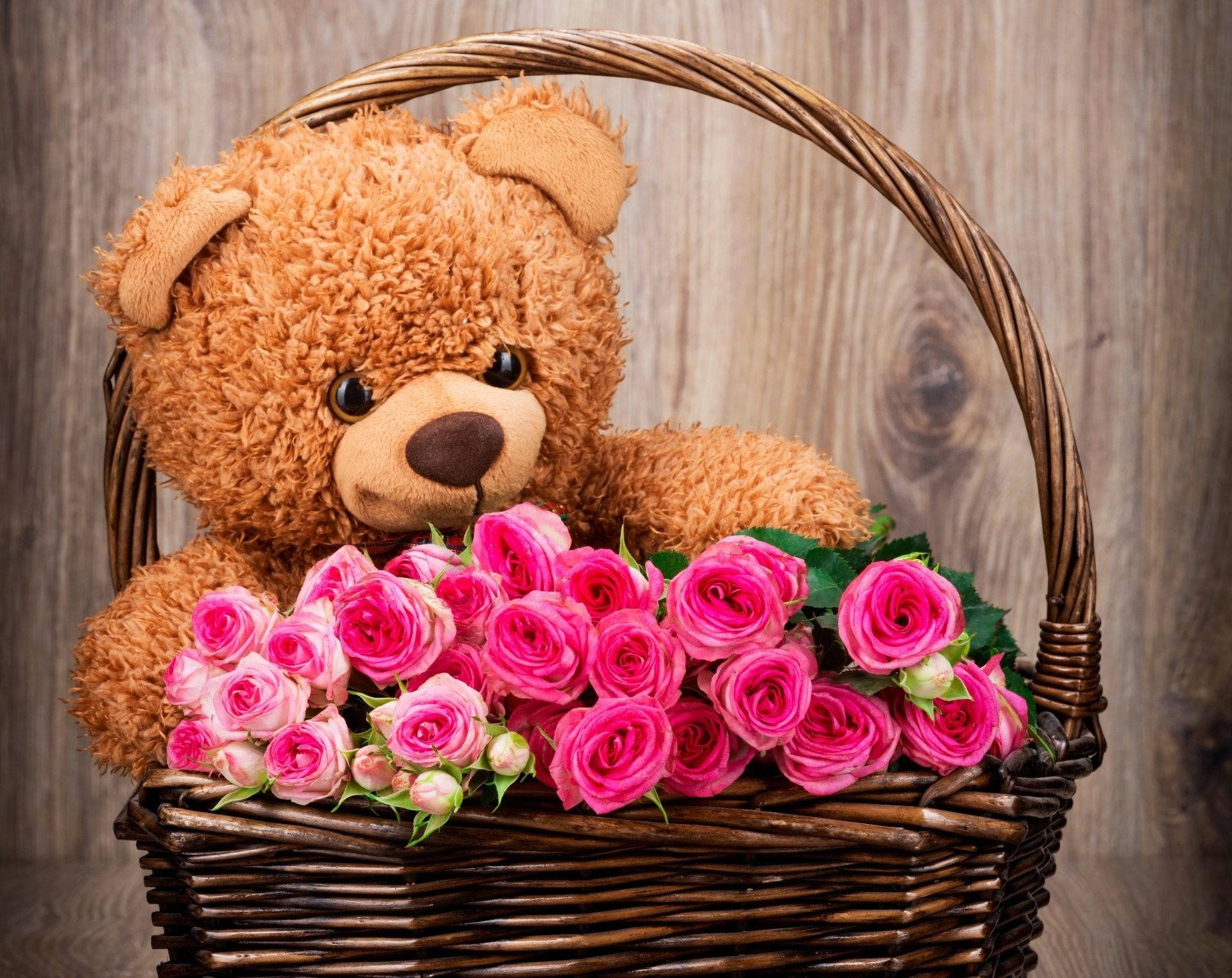 Teddy Bear 桌布 壁紙 — 一隻毛茸茸的棕色泰迪熊坐在柳條籃中，籃子裡裝滿了鮮豔的粉紅玫瑰，背景是質樸的木頭。