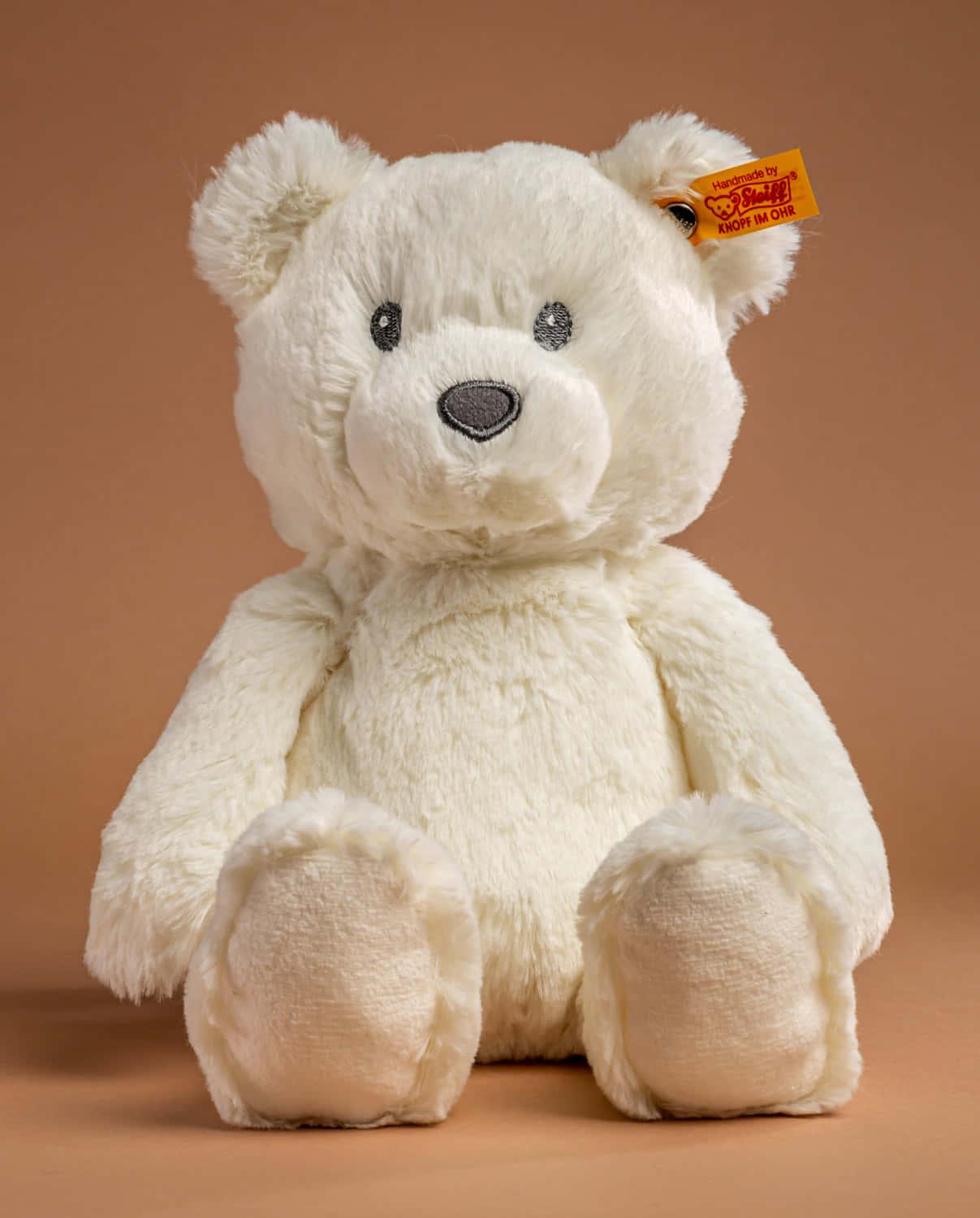 Cuddly 桌布 壁紙 — 一隻毛茸茸的白色Steiff泰迪熊直立坐在溫暖的棕色背景前，帶有可見的品牌標籤。