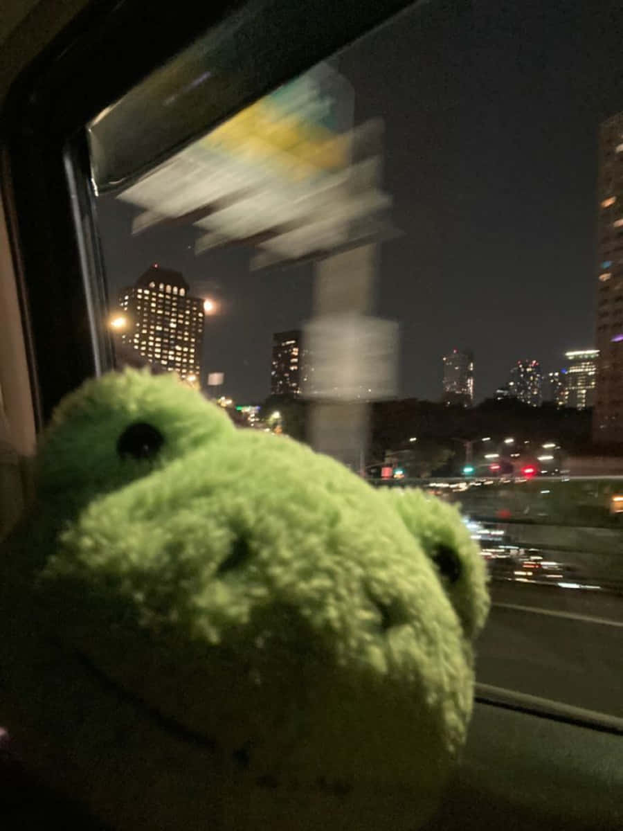 Aesthetic Frog 桌布 壁紙 — 一隻毛茸茸的綠色絨毛青蛙從火車窗戶探出頭來，夜晚模糊的城市燈光呼嘯而過。