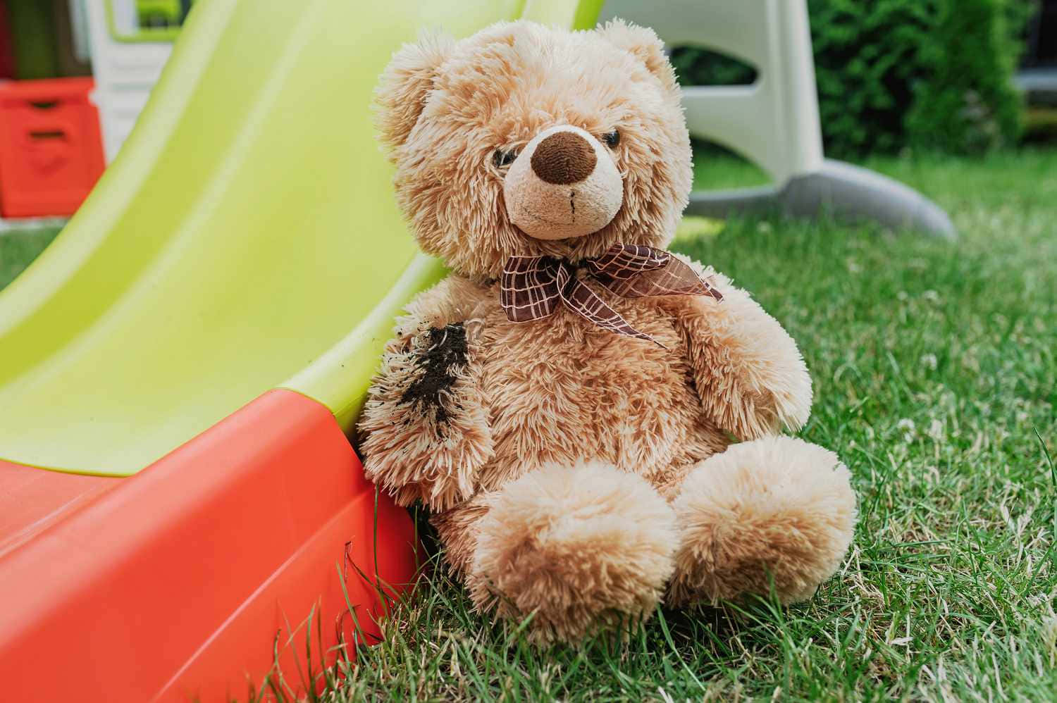 Teddy Bear 桌布 壁紙 — 一隻沾滿泥巴的棕色泰迪熊戴著格子蝴蝶結，坐在綠色草地上，旁邊是一個鮮黃色和紅色的塑膠溜滑梯。