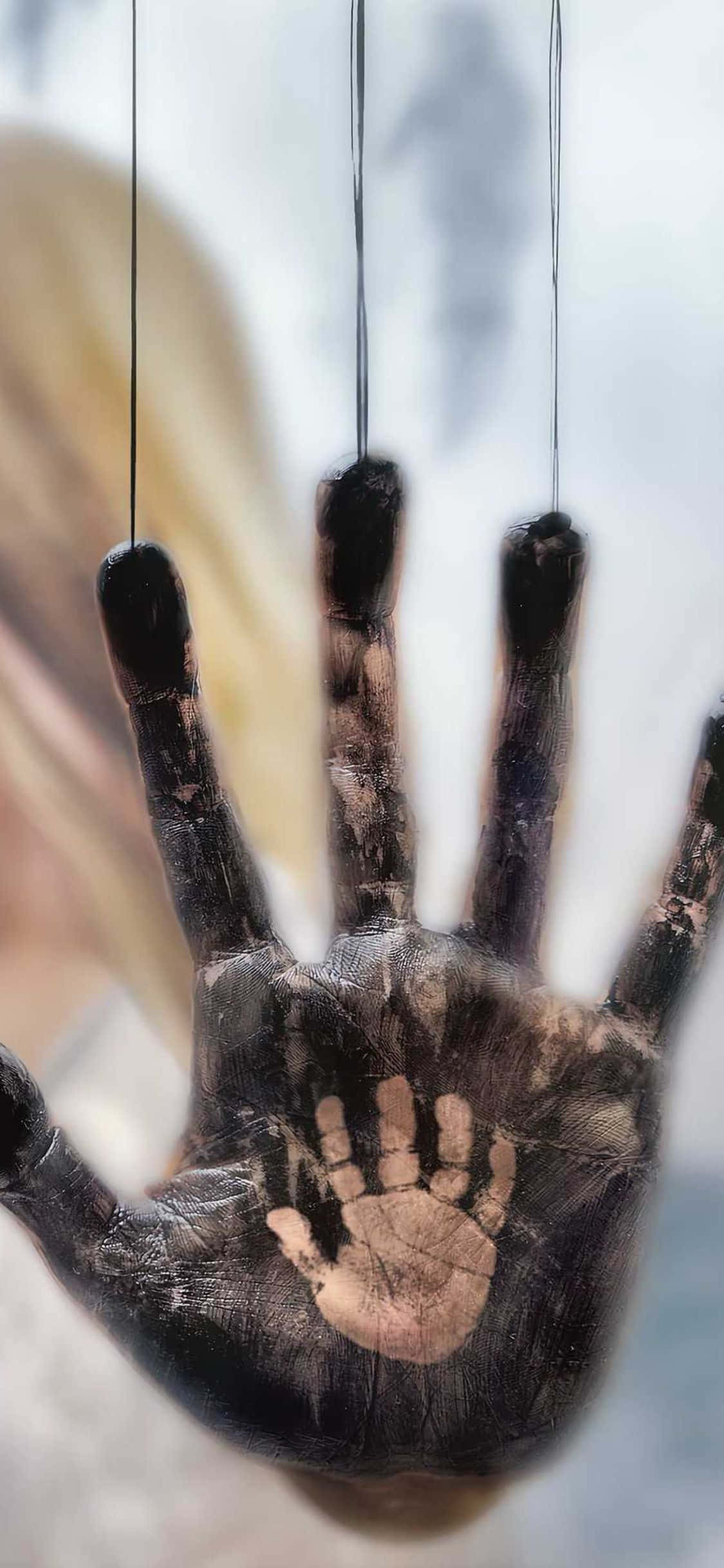 iPhone X Death Stranding 桌布 壁紙 — 一隻沾滿深色油漆的手，用細線懸掛在模糊背景前的特寫。